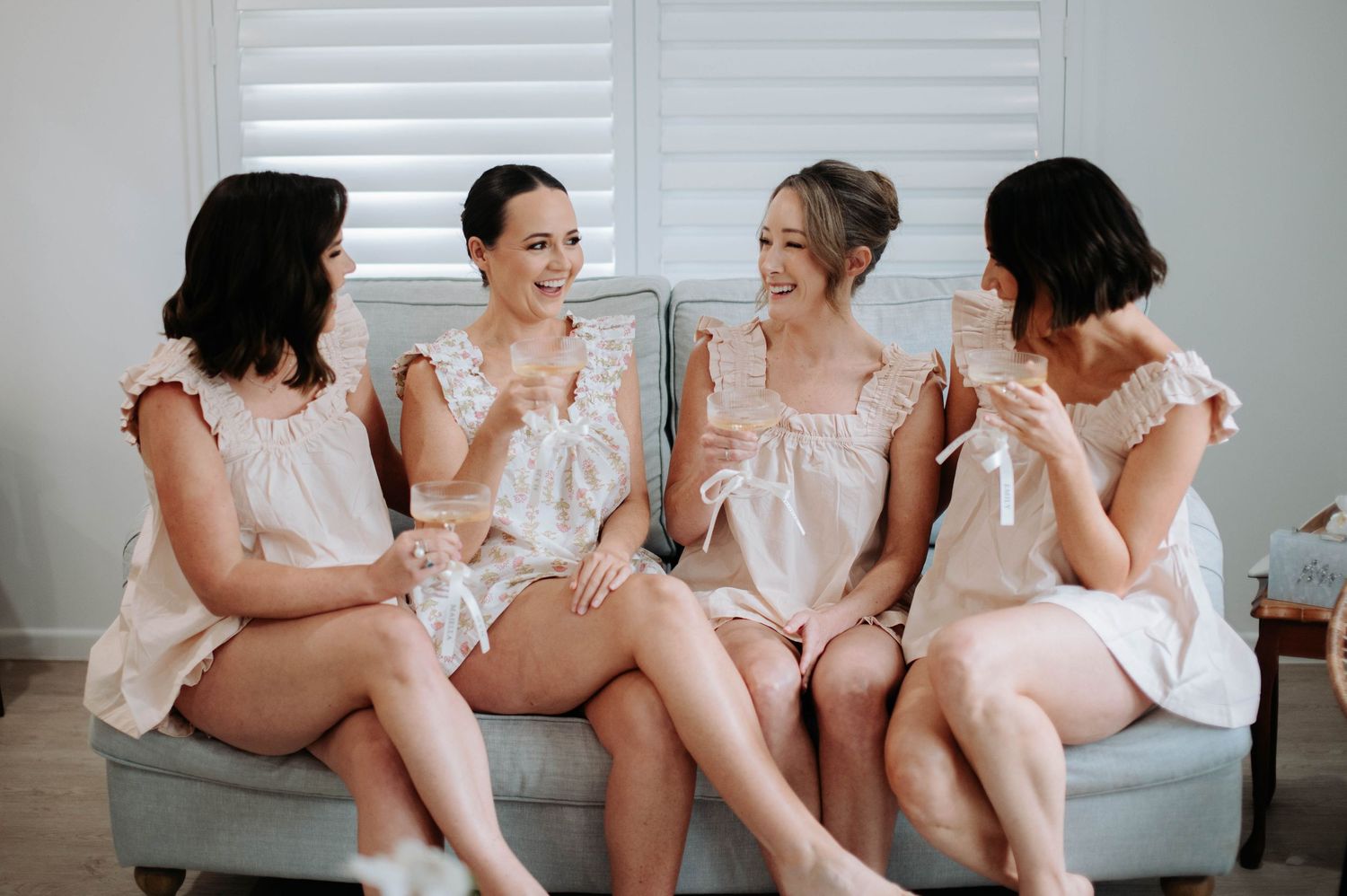 Four women in blush pink dresses sit together on a grey sofa in a bright interior setting.