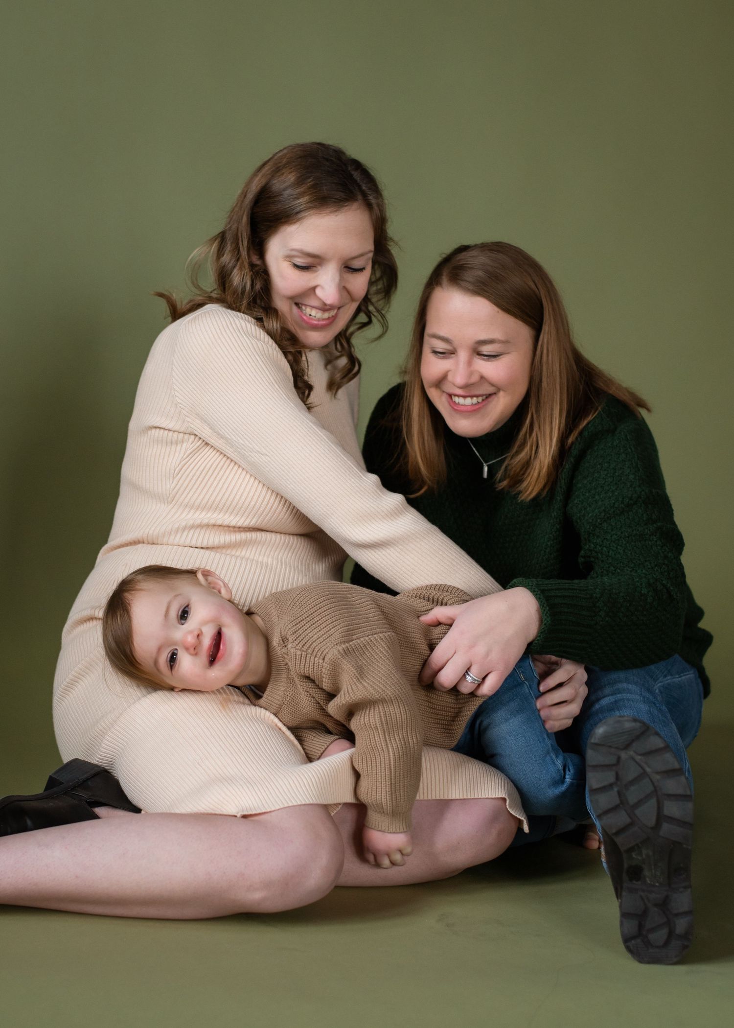 A toddler in a brown sweater sits with a woman in a beige sweater dress on a plush carpet by a leather couch.