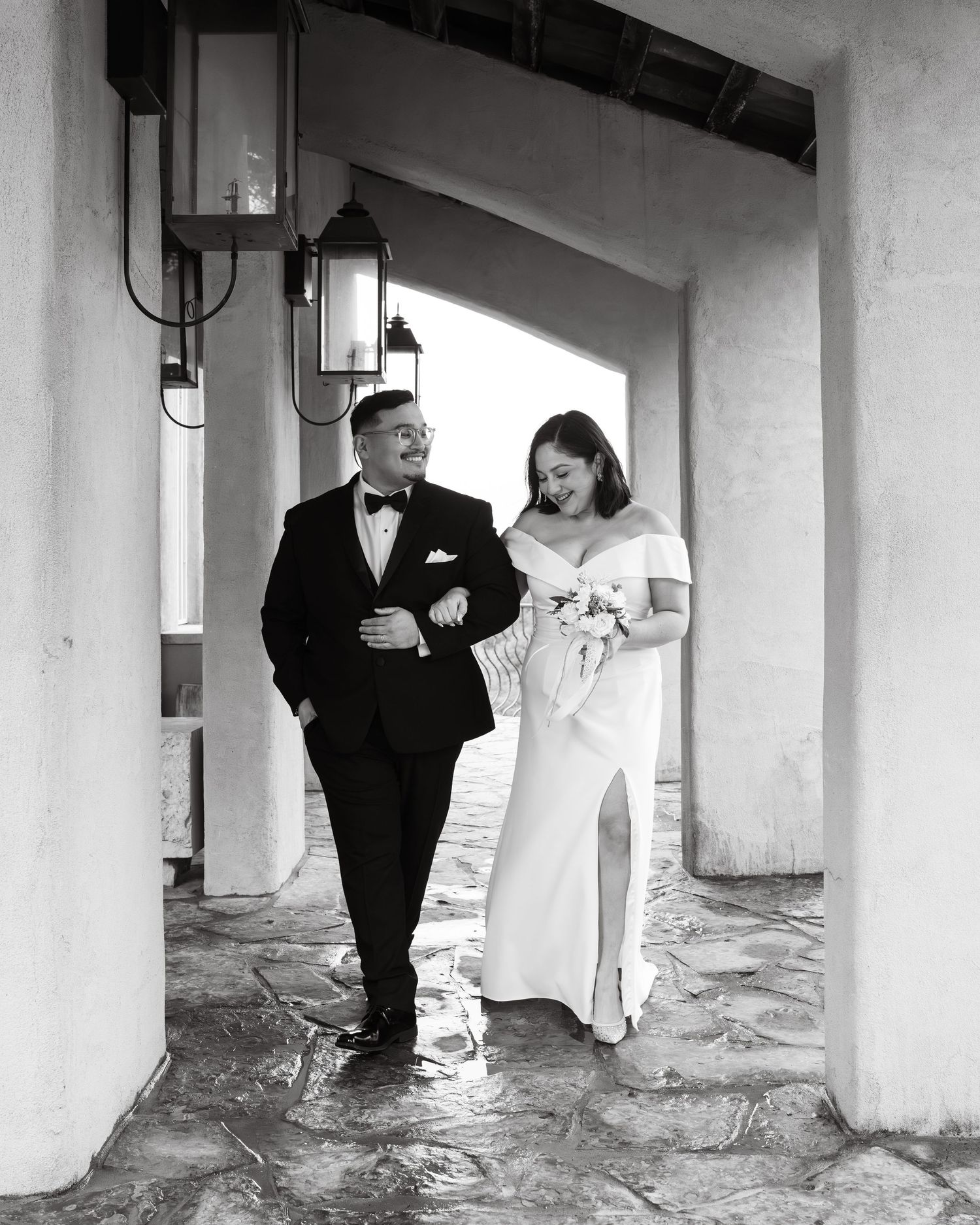 Couple in formal attire walks through covered walkway in elegant black and white photo.