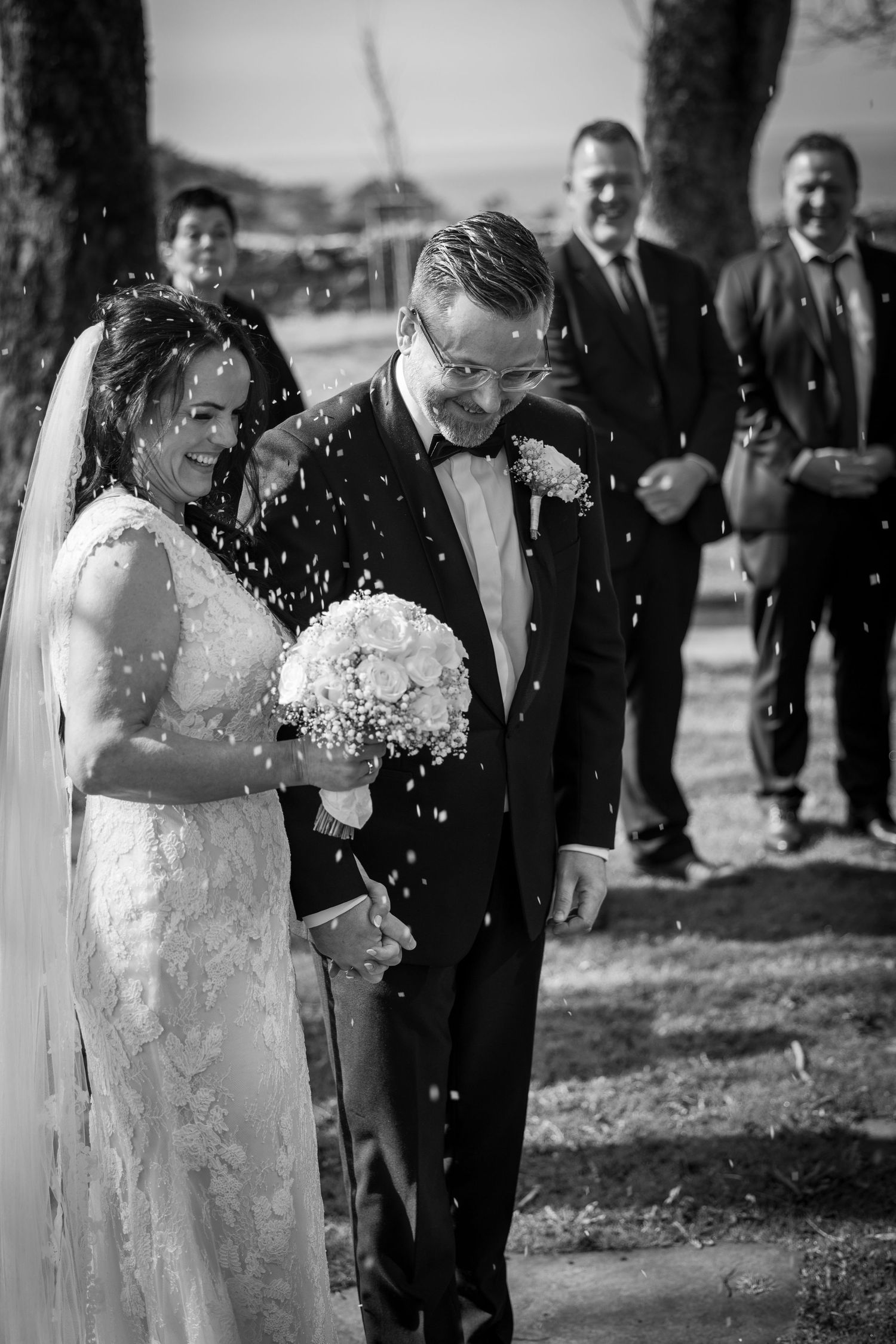 Bride and groom walk through confetti shower outdoors, guests celebrating behind them in black and white photo.