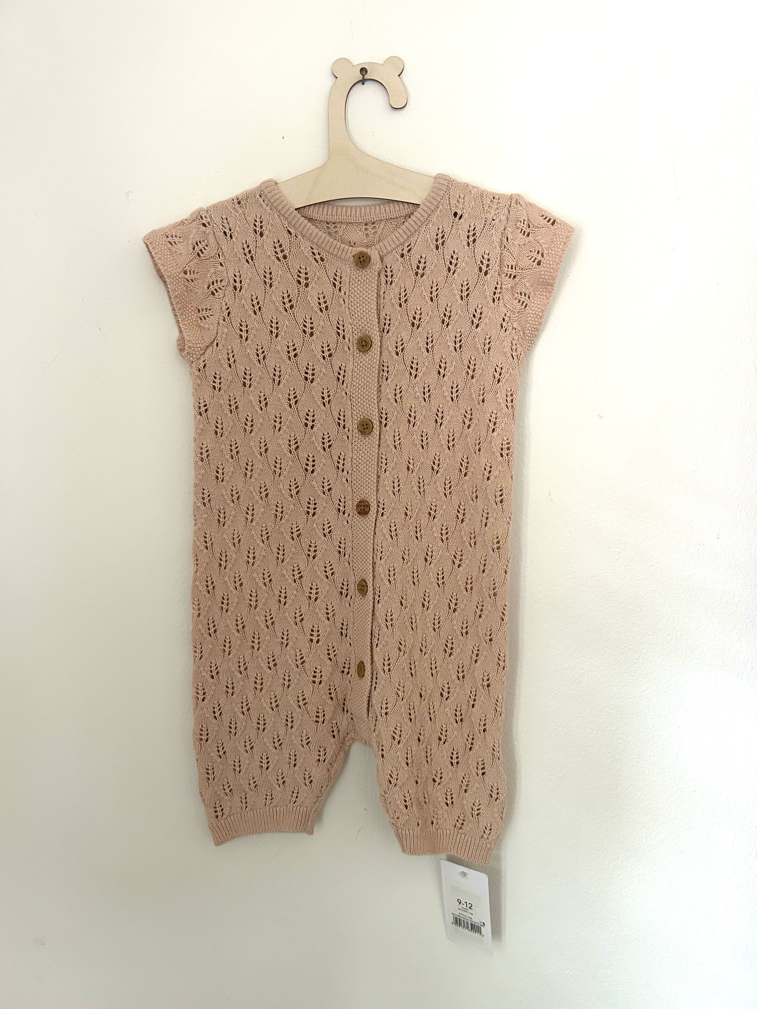 Vintage-style beige knitted baby romper with button details and side pockets hanging on display.