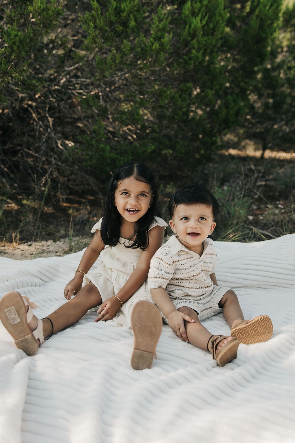 Two young children sitting together on a white blanket outdoors wearing casual clothing and sandals.