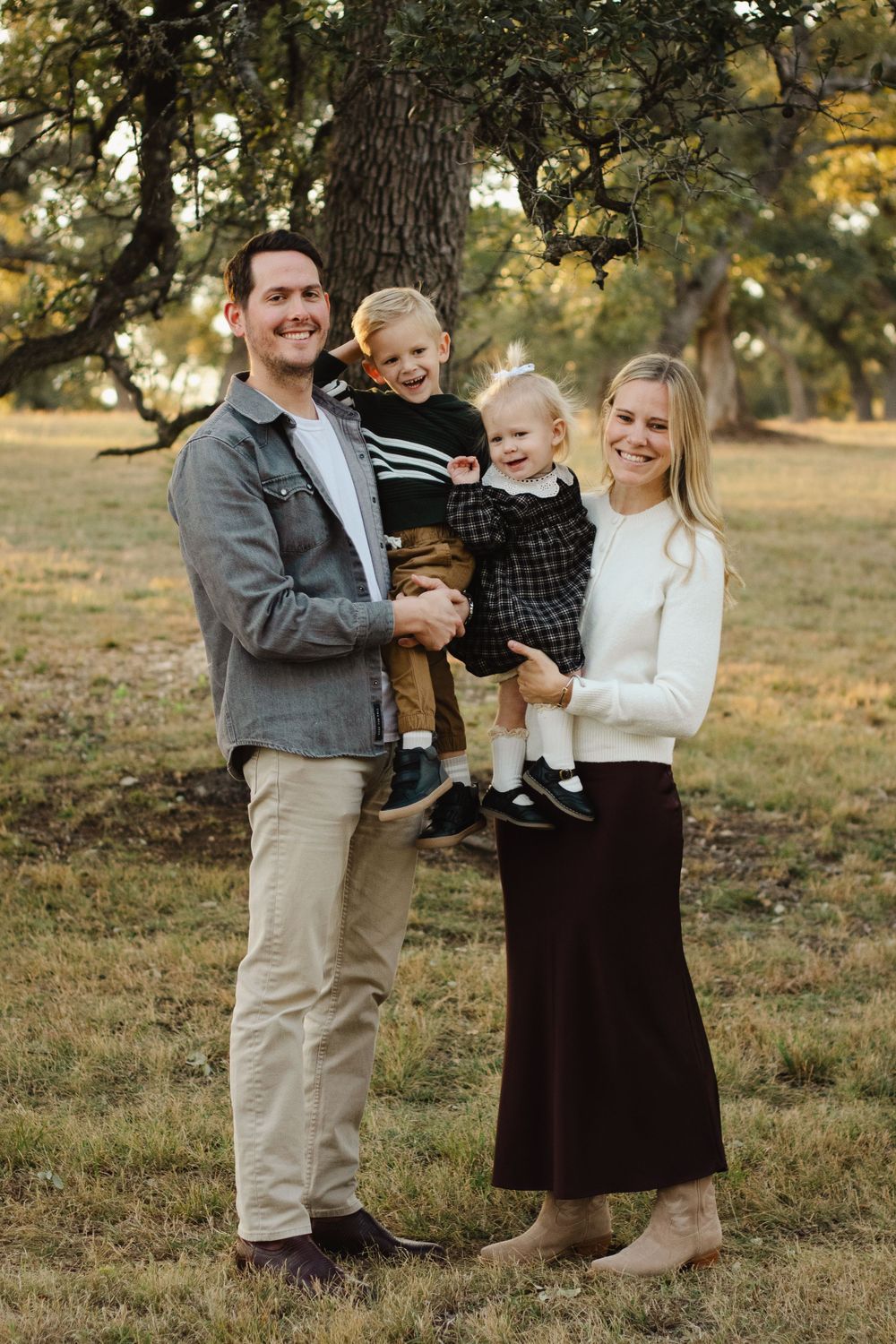 Family portrait in casual attire standing together in a grassy field with oak trees.