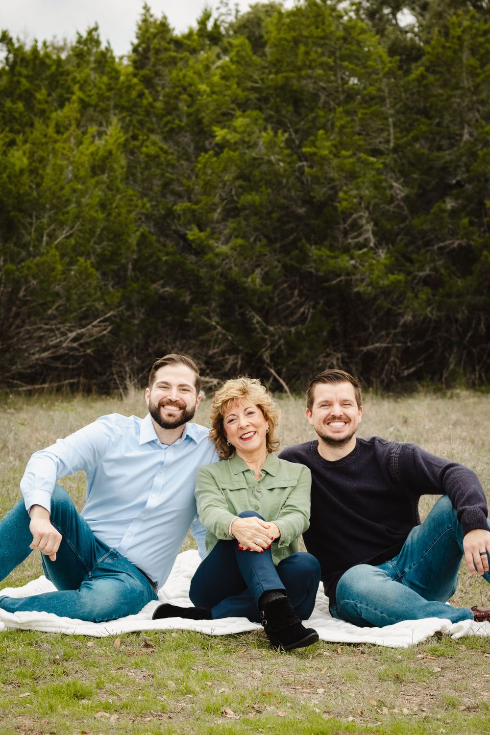 Three people sitting together on a blanket outdoors with evergreen trees in the background.