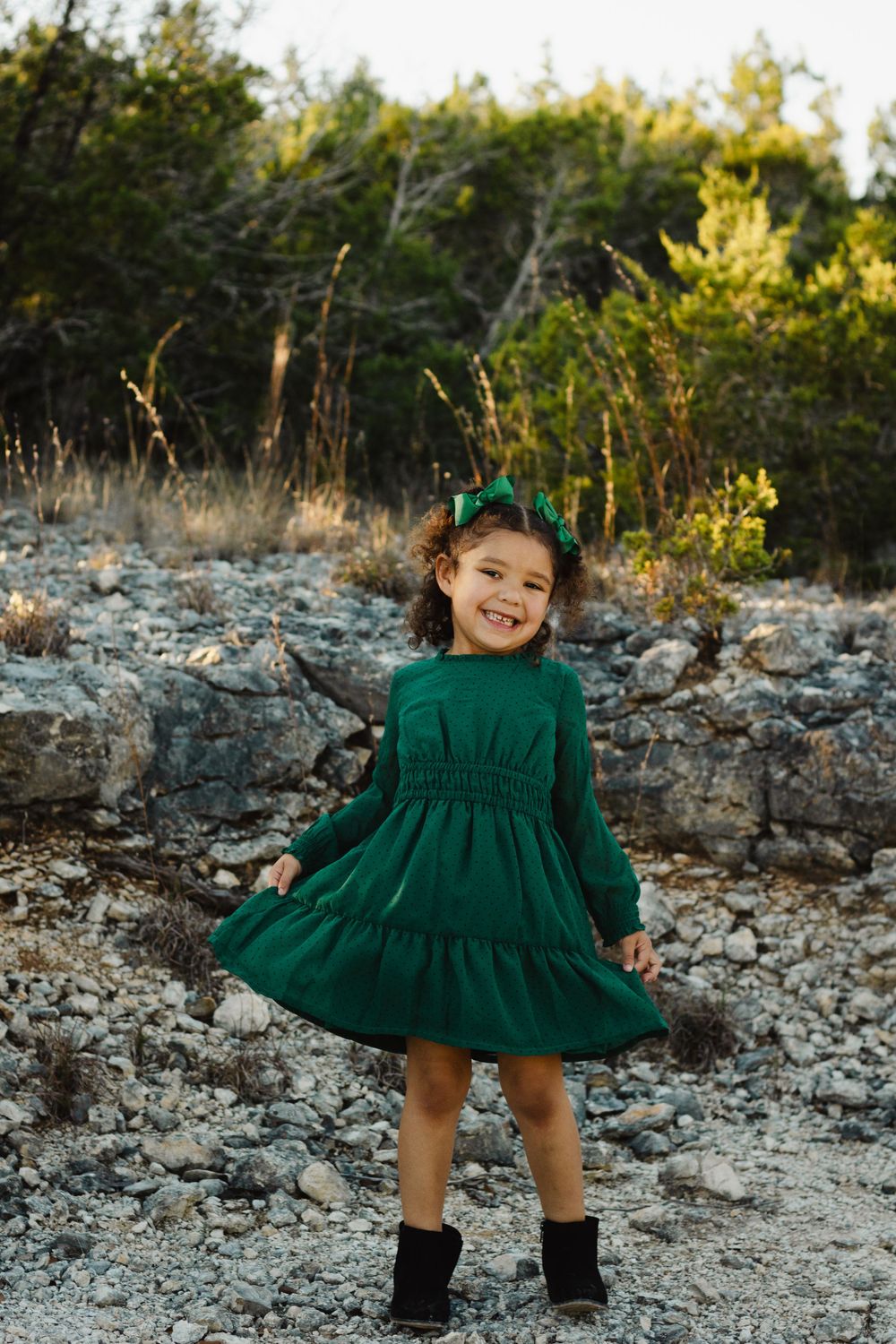 Child in green dress twirling and smiling on rocky terrain with trees in background.