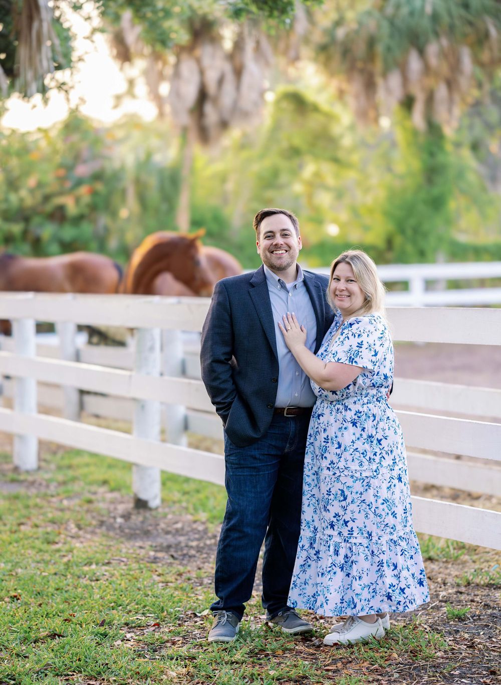 Happy couple in formal attire pose together by a white fence with horses visible in the background at a rustic farm setting.