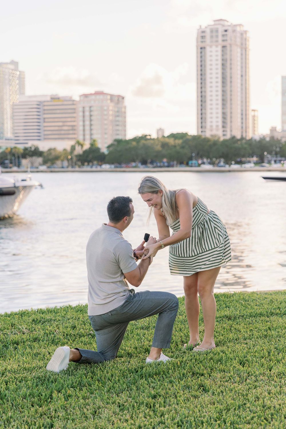 Man proposes on one knee to woman by waterfront with city skyline, she reacts with joy to engagement ring.