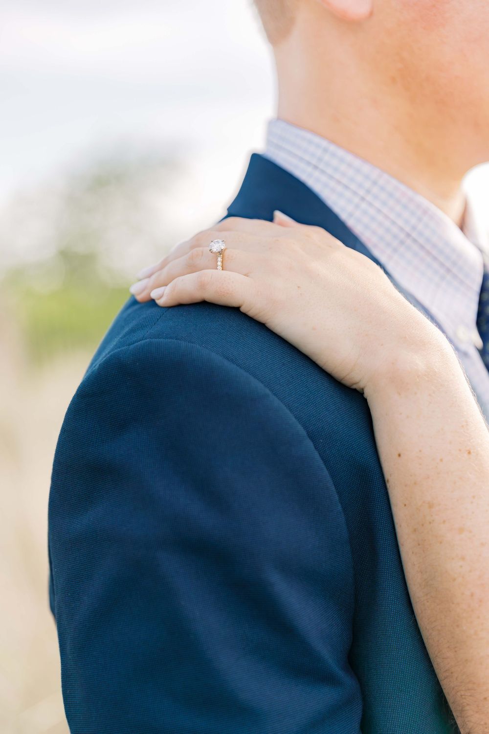 Close up of engagement ring as woman's hand rests on groom's navy blue suit jacket shoulder.