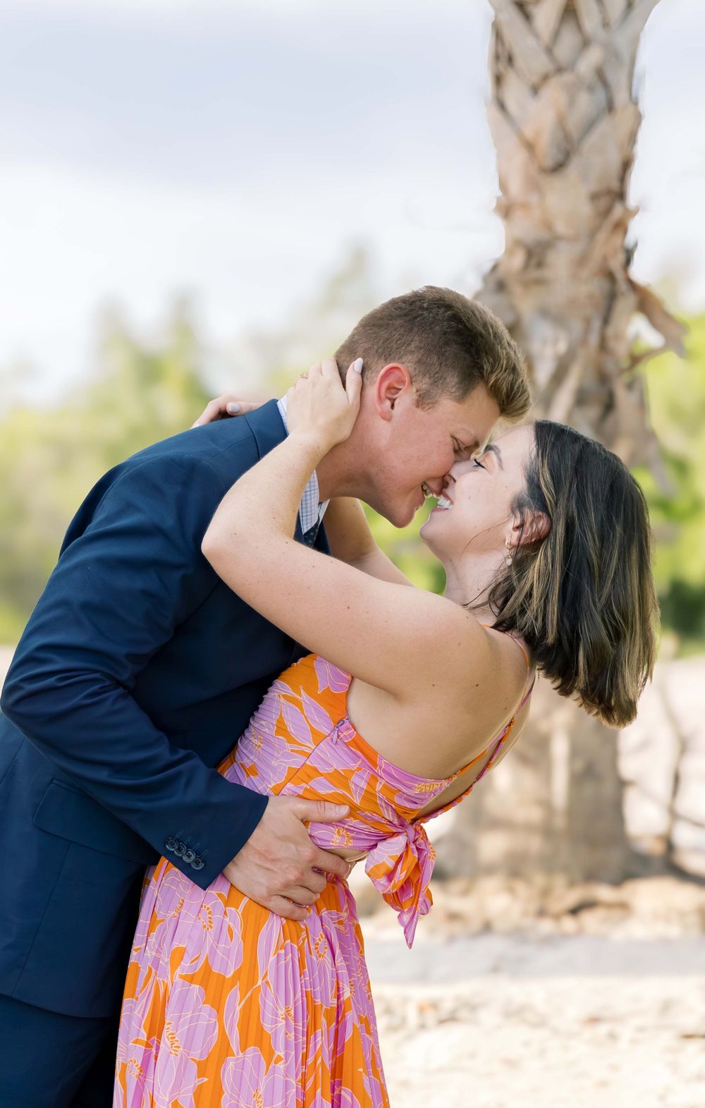 A romantic couple embraces outdoors, with the woman wearing a pink floral sundress and the man in a navy suit.