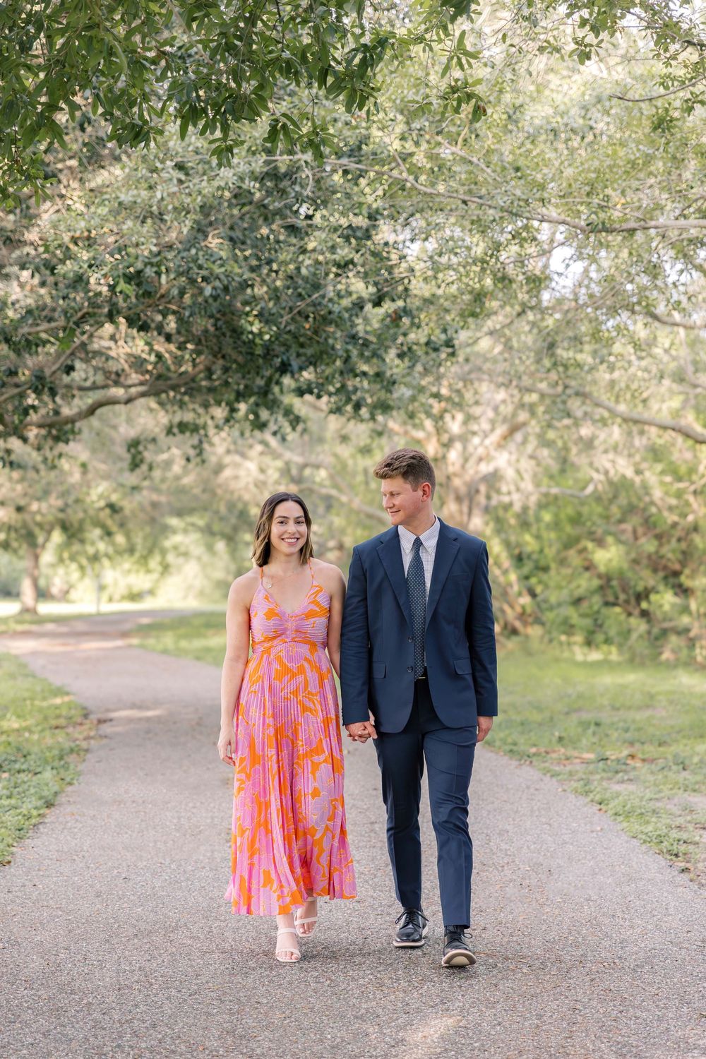 A couple strolls together on a garden path, with her wearing a vibrant orange dress and him in a navy suit.