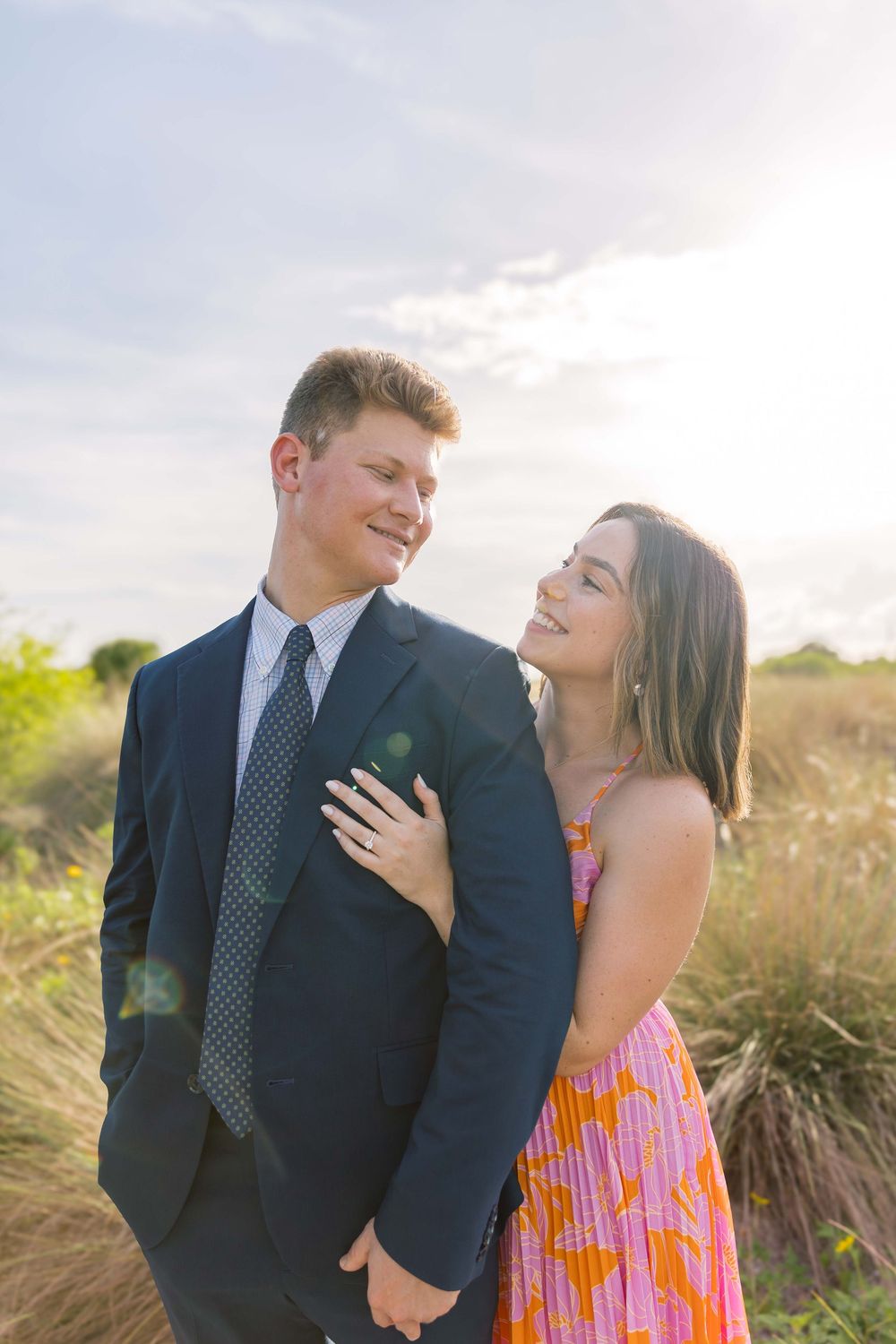 Happy couple embraces in a field at sunset, with her in a floral sundress and him in a navy suit and tie.