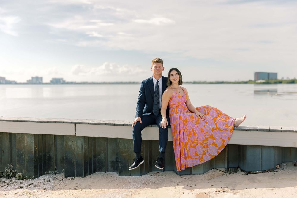A couple sits together on a seawall at sunset, with the woman wearing a bright floral dress by the waterfront.