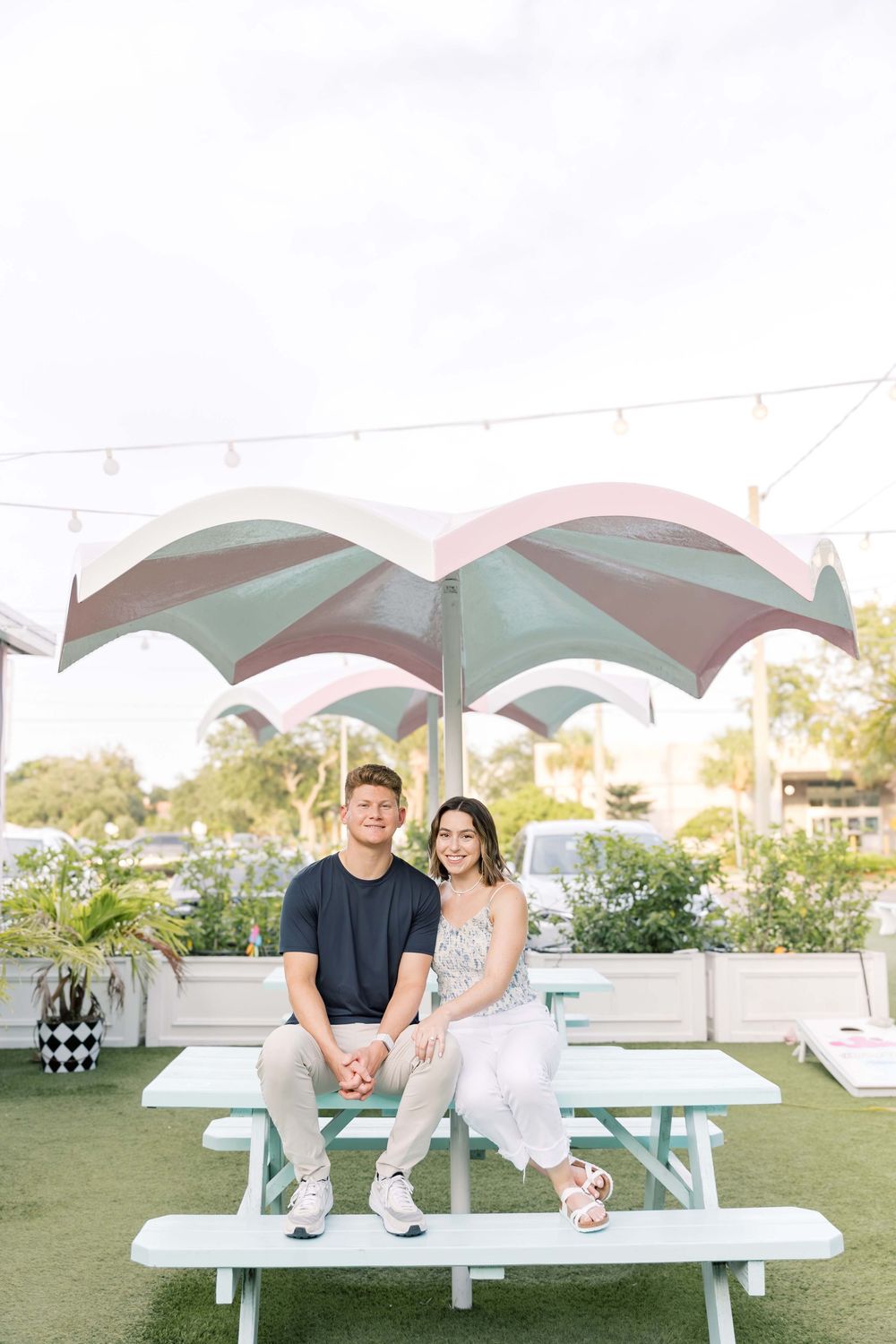A couple sits together under a striped umbrella at a picnic table on an outdoor patio space with greenery.