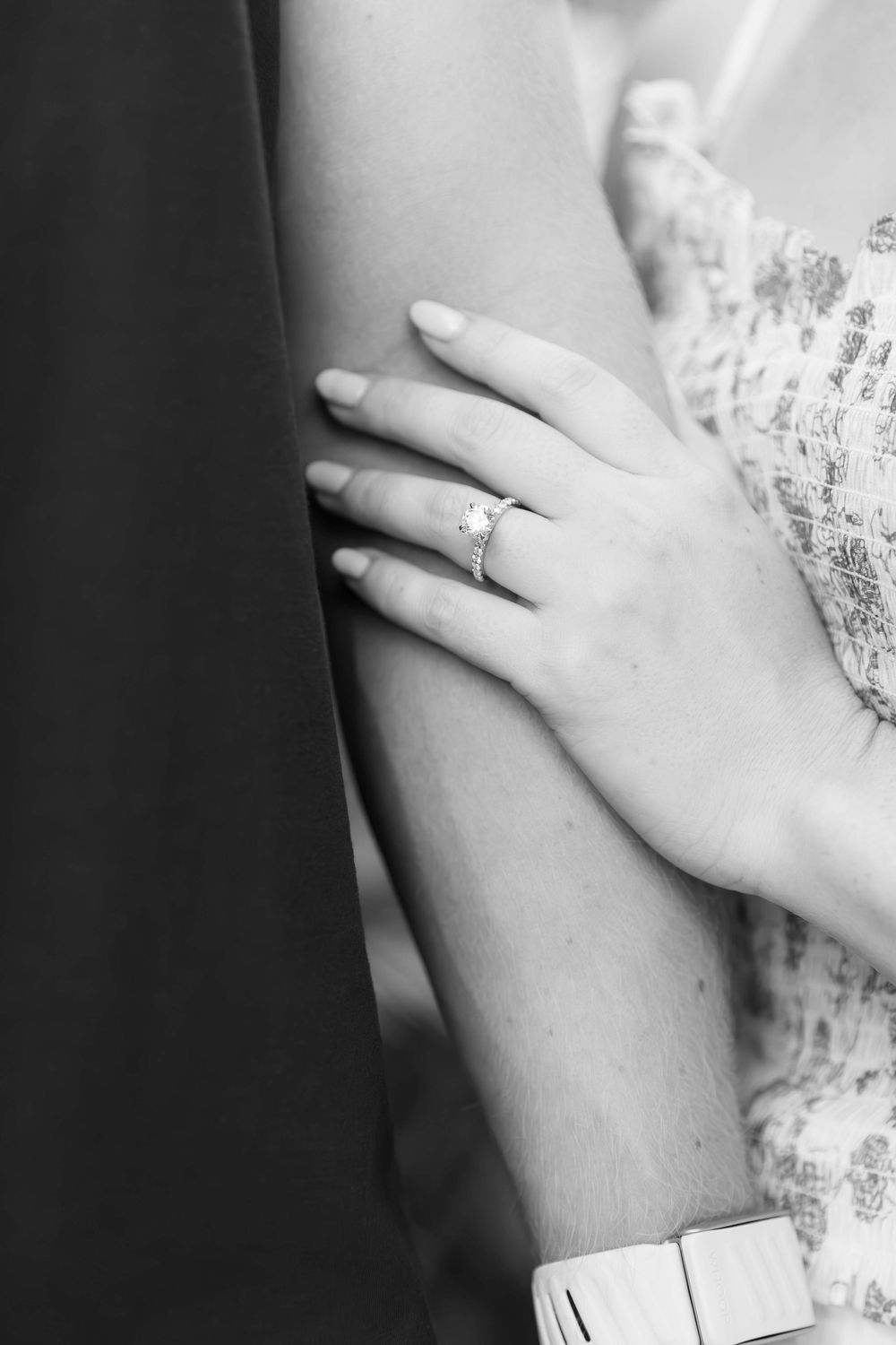 Black and white close-up shot of a hand wearing an engagement ring resting against a flowered dress sleeve.