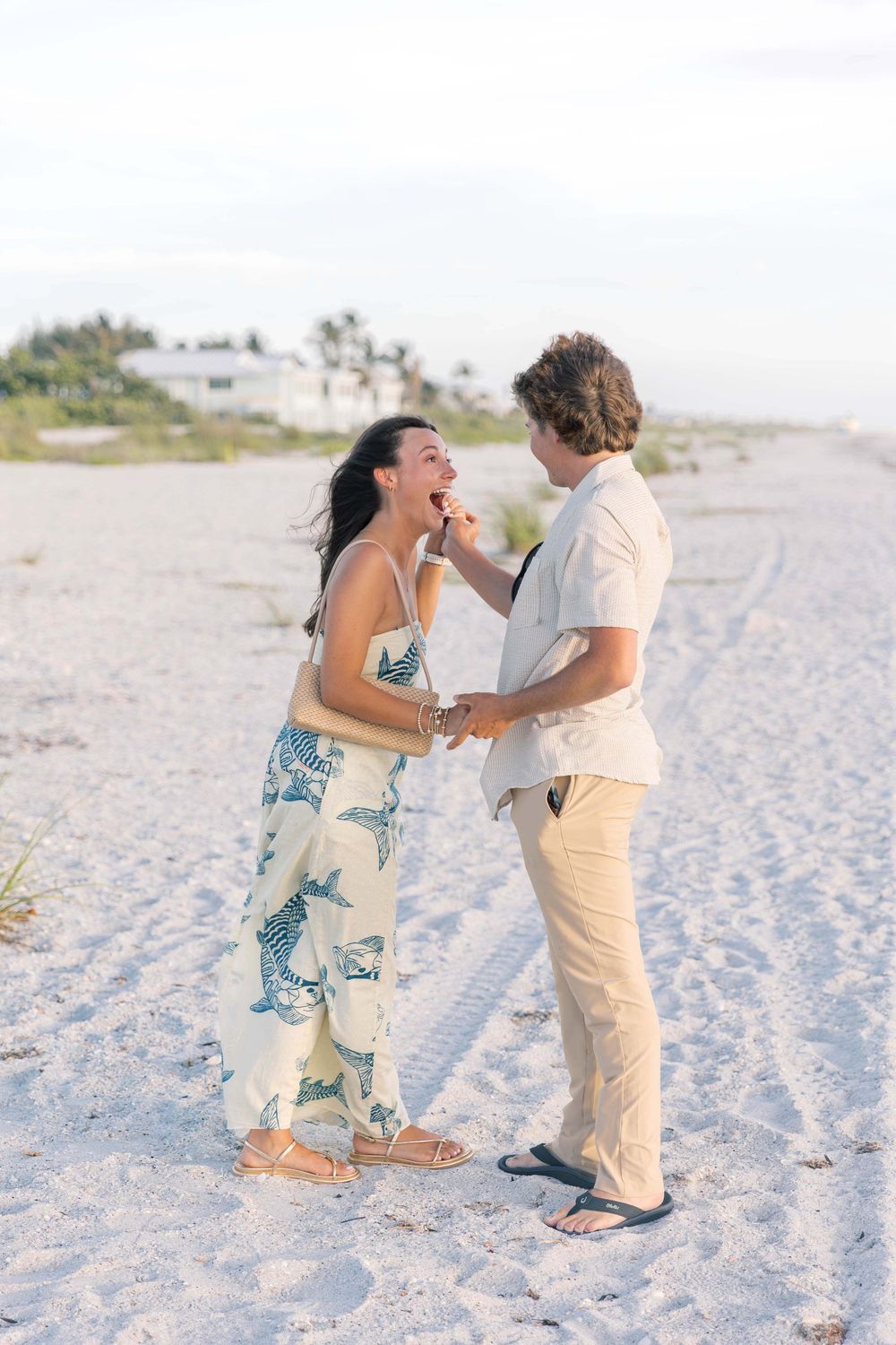 Couple sharing a romantic moment on a sandy beach, woman in floral jumpsuit laughing as man feeds her a treat.