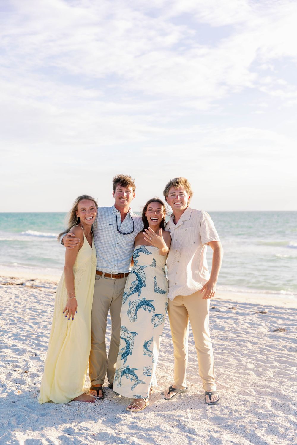 Four smiling young adults pose together on a white sandy beach with turquoise ocean waves in the background.