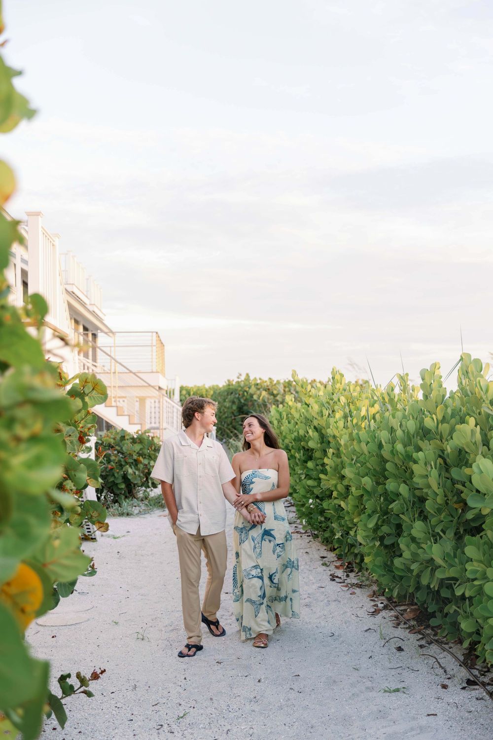 Couple walking hand in hand along sandy beach path lined with lush green foliage near coastal buildings.
