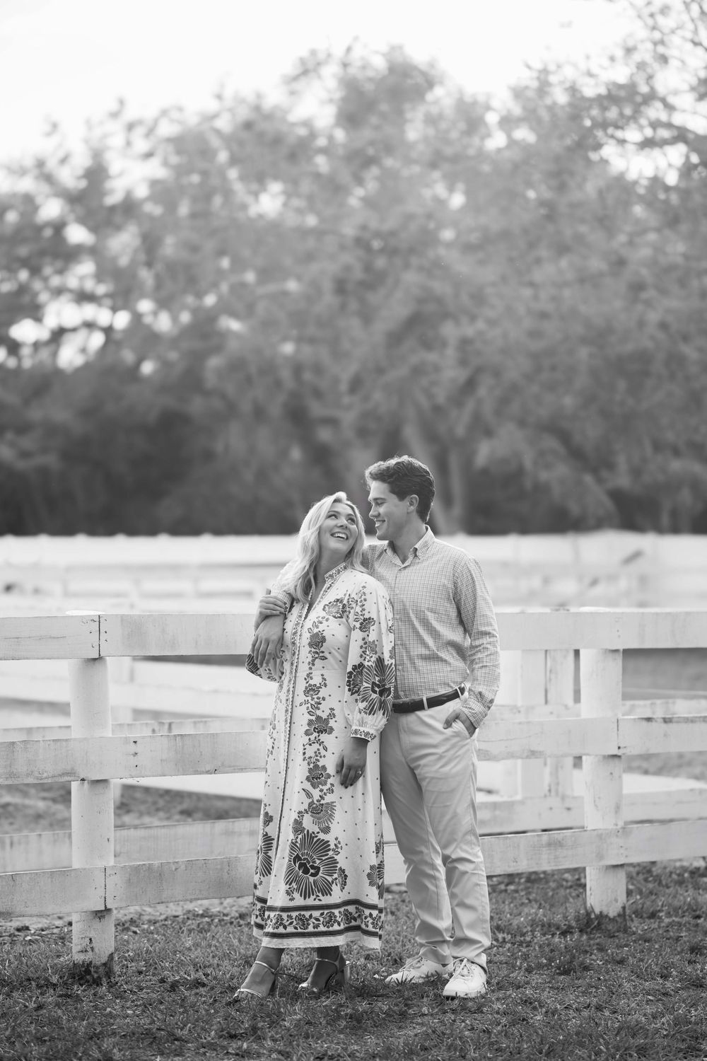 A romantic black and white photo of a couple leaning against a white wooden fence in a pastoral countryside setting.