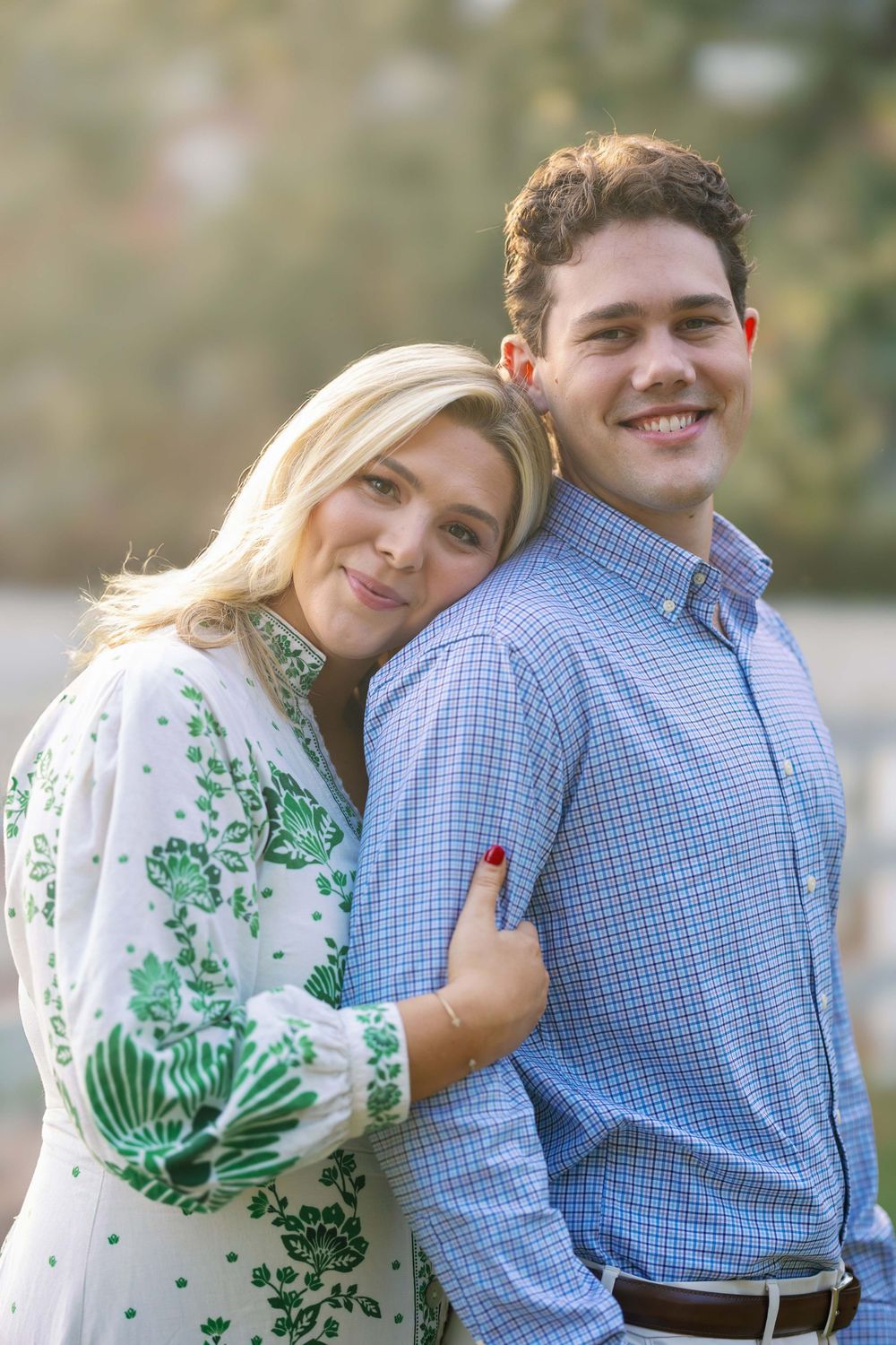 A couple poses together outdoors, wearing a blue checkered shirt and white dress with green floral pattern.