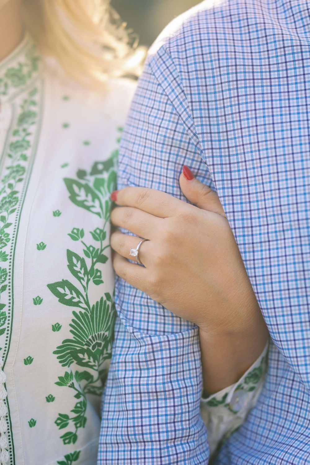 Close up shot of a diamond engagement ring on hand clutching a blue checkered shirt against a floral print background.