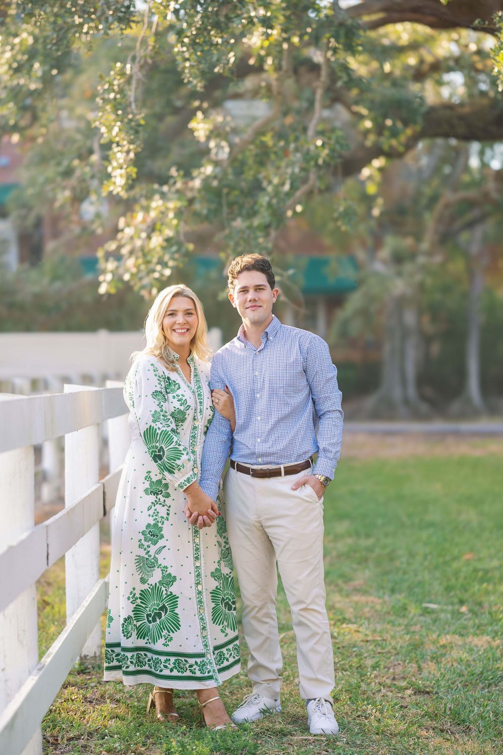 A couple poses by a white fence in a garden setting, with the woman wearing a green and white patterned dress.