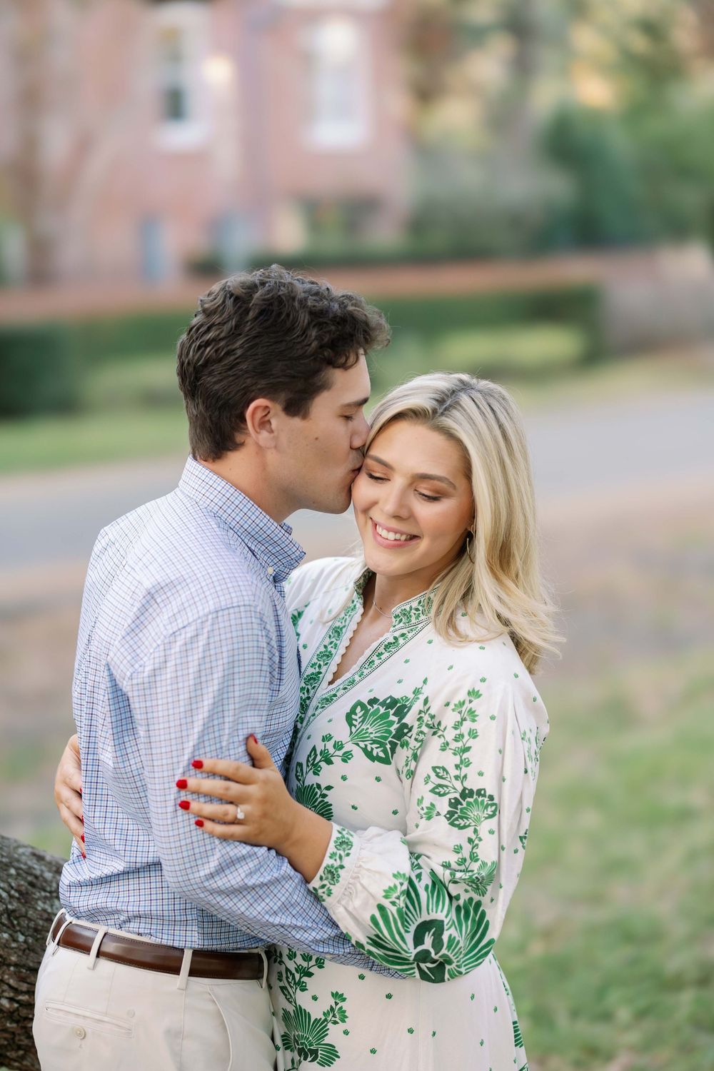 A romantic outdoor portrait of a couple embracing, with her wearing a white and green floral dress near a brick building.
