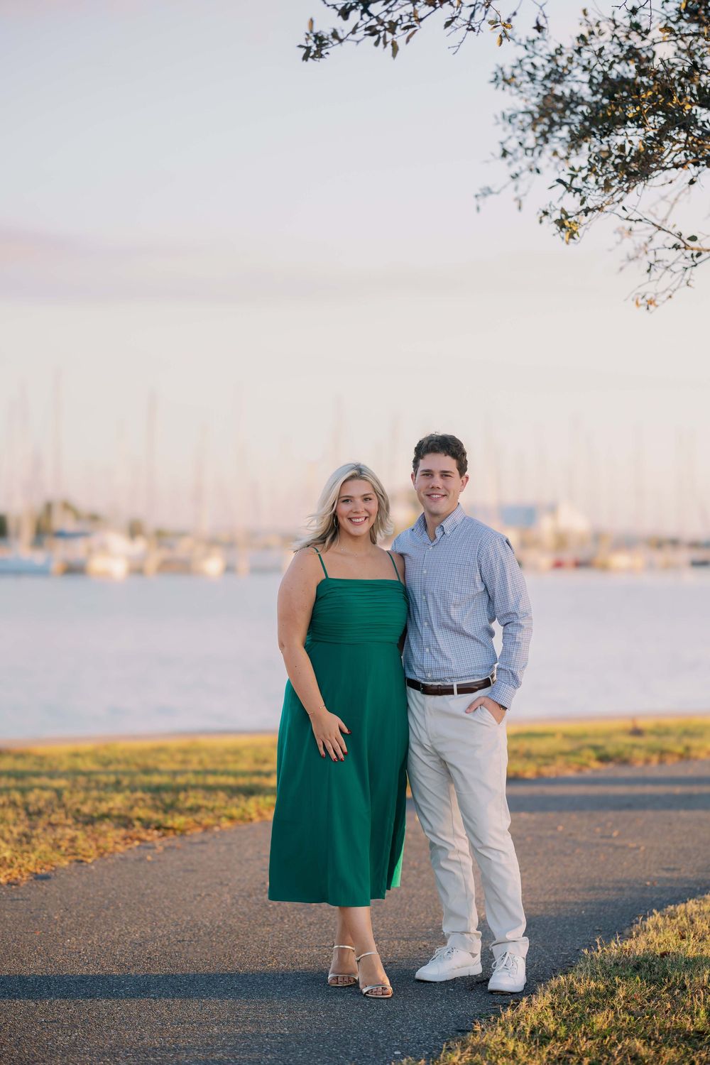 A couple poses together on a waterfront path at sunset with boats and a marina visible in the background.