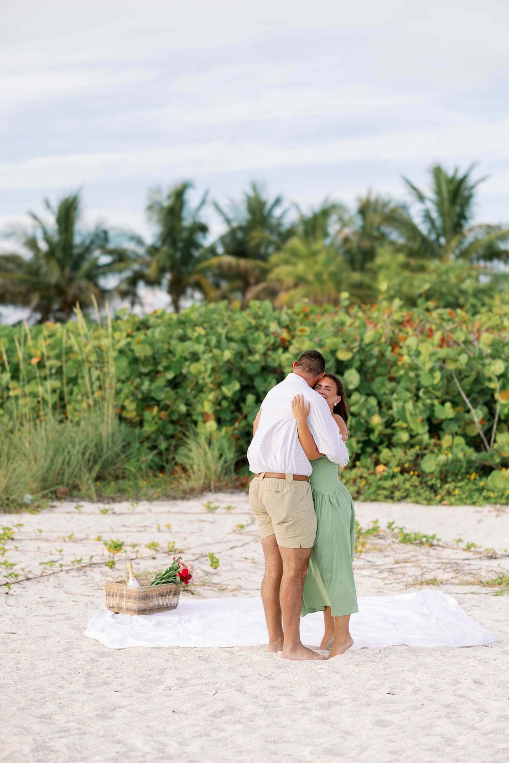 Couple embracing on sandy beach with tropical greenery, picnic basket nearby, romantic engagement moment outdoors.