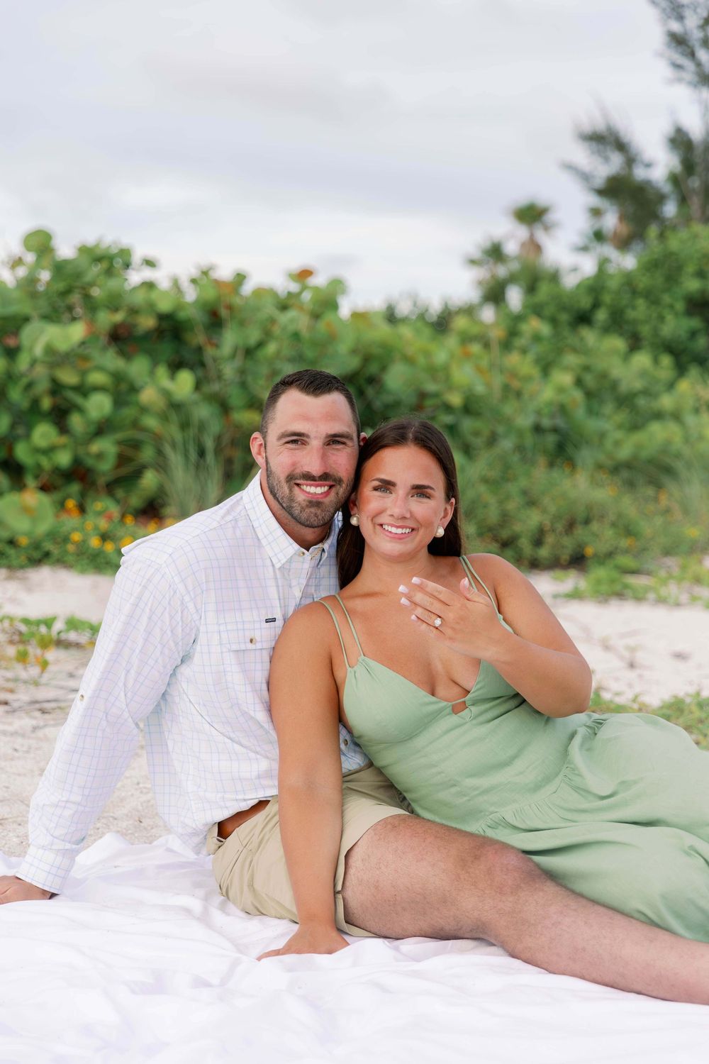 Happy couple sitting on white blanket at sandy beach, woman in green dress showing engagement ring, smiling together.