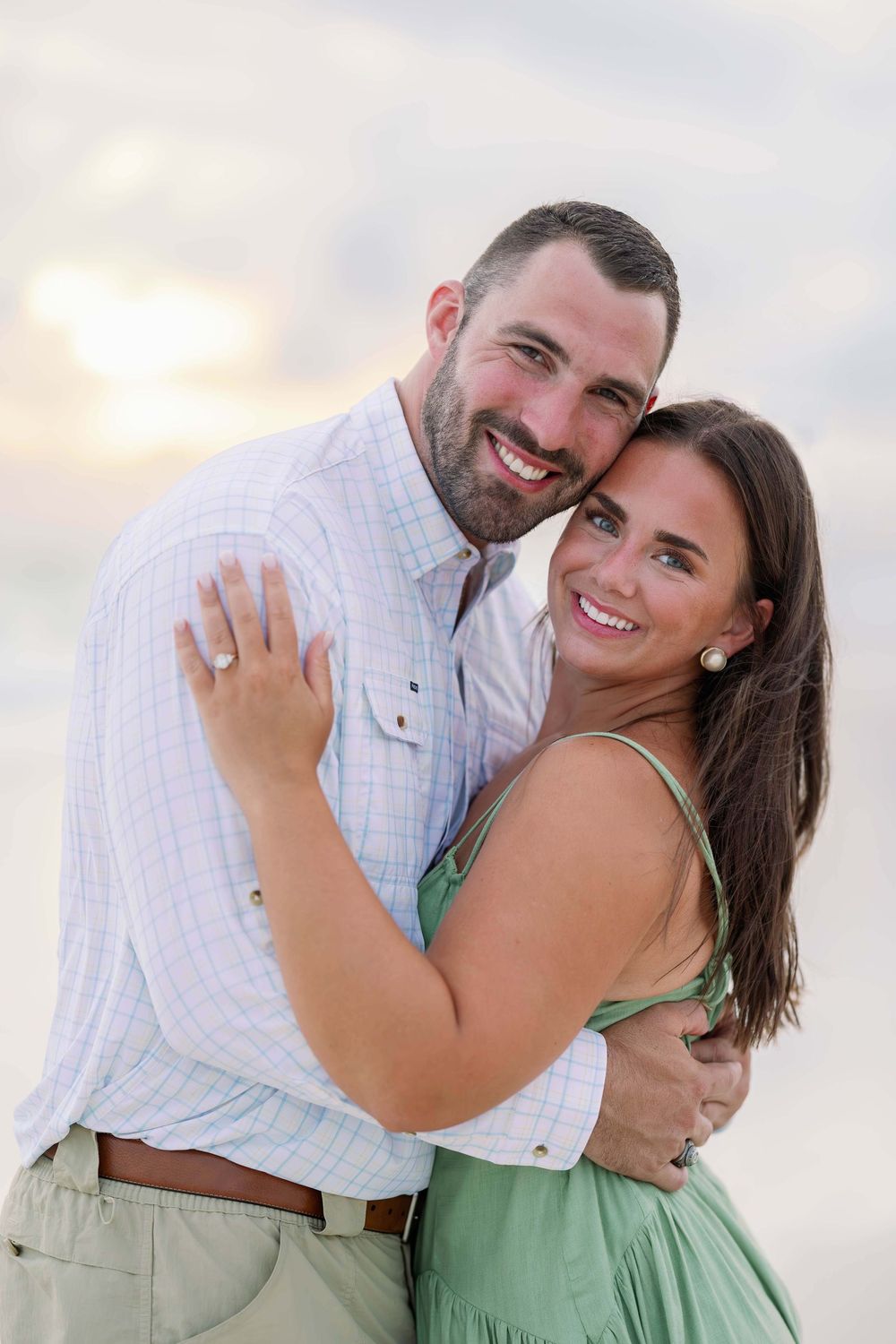 Happy couple embracing on a beach at sunset, woman in green dress with engagement ring, man in light blue shirt smiling.