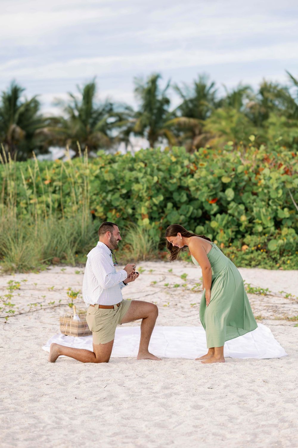 Man proposes on one knee to woman in green dress on white sand beach with tropical palms backdrop.
