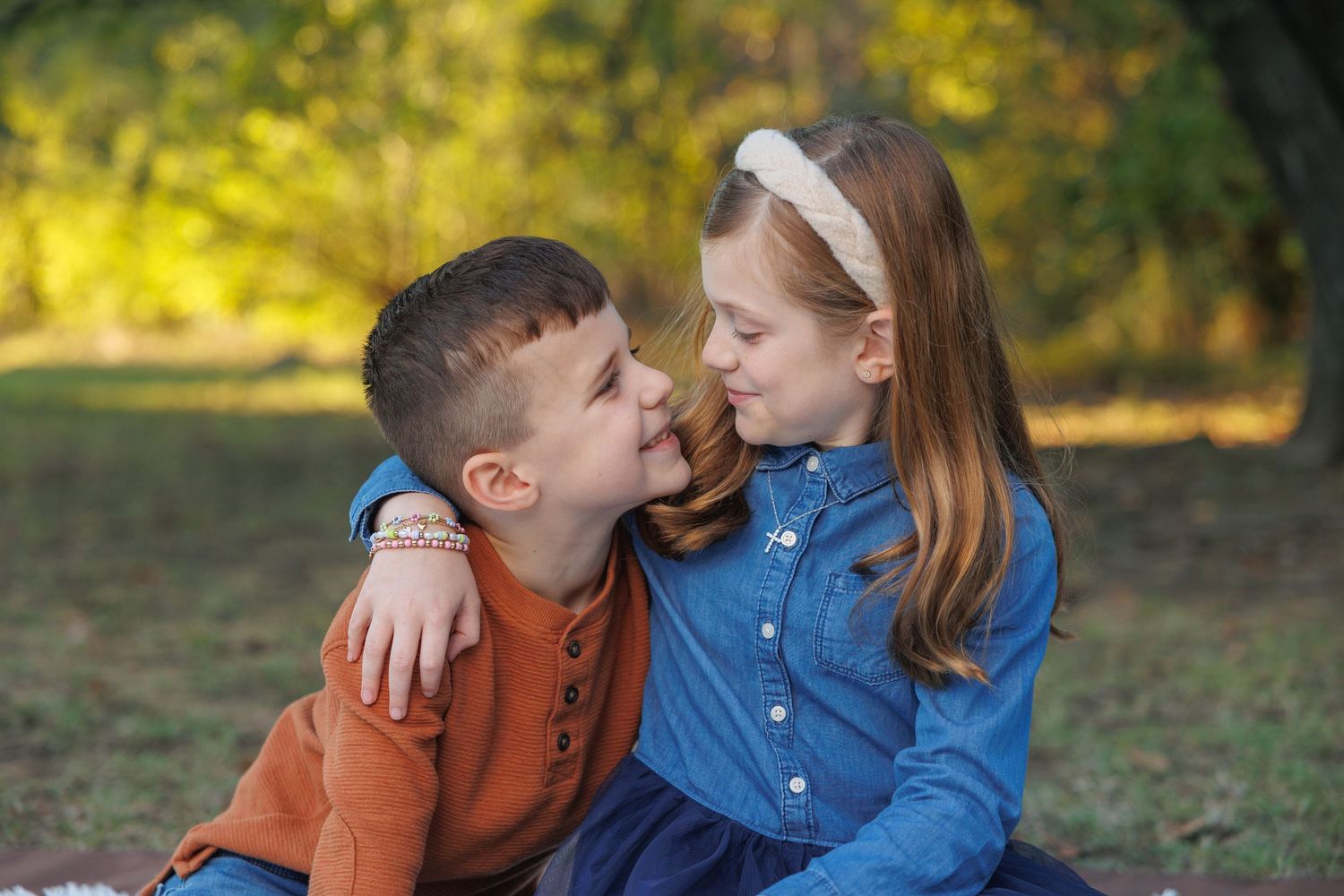 Two young children share a sweet moment together while sitting outdoors in autumn scenery.