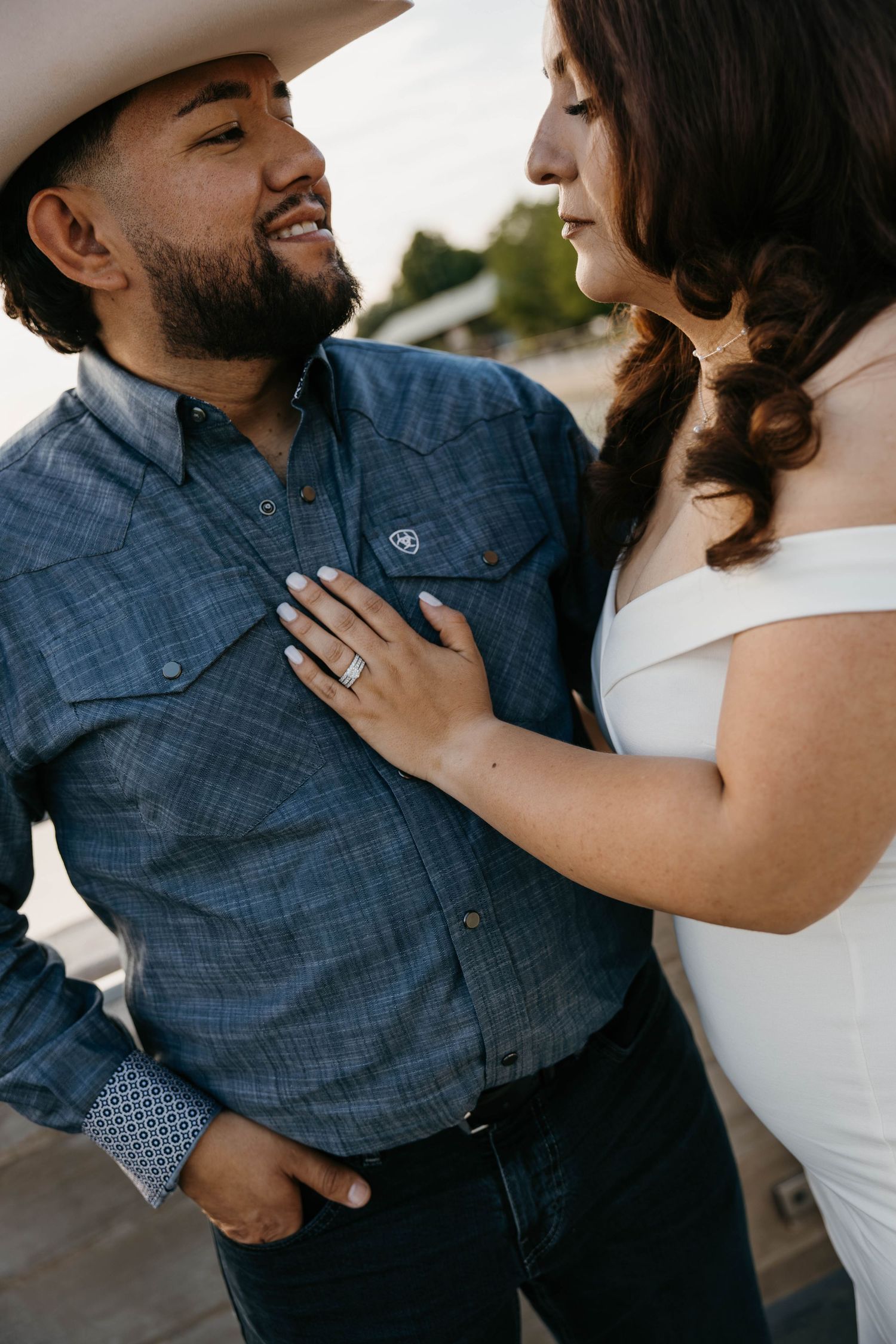 Smiling groom in cowboy hat and blue Ariat shirt gazes at bride during outdoor engagement session.