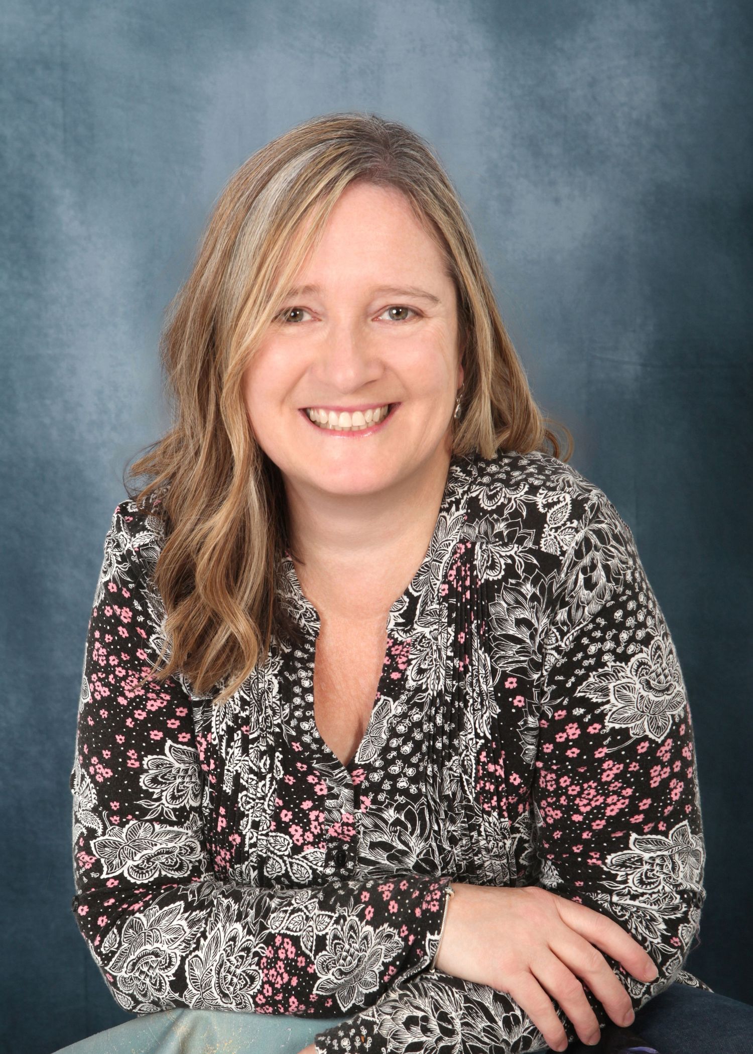 Professional portrait of someone in a patterned black and pink floral blouse against a mottled blue backdrop.