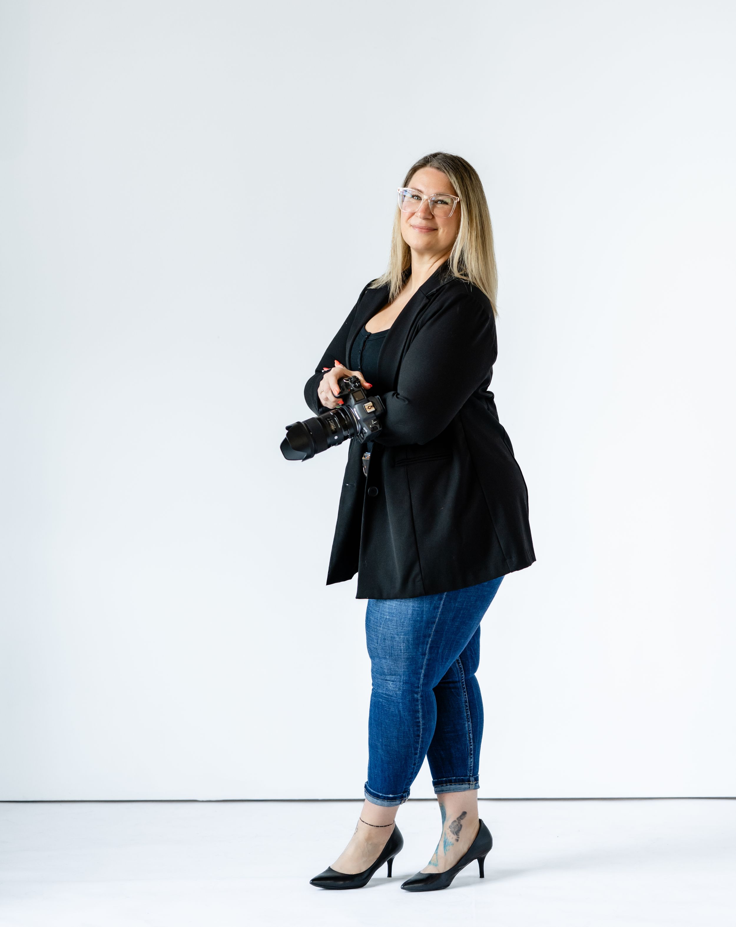 Professional photographer in a black blazer, blue jeans and heels stands against white background holding camera.