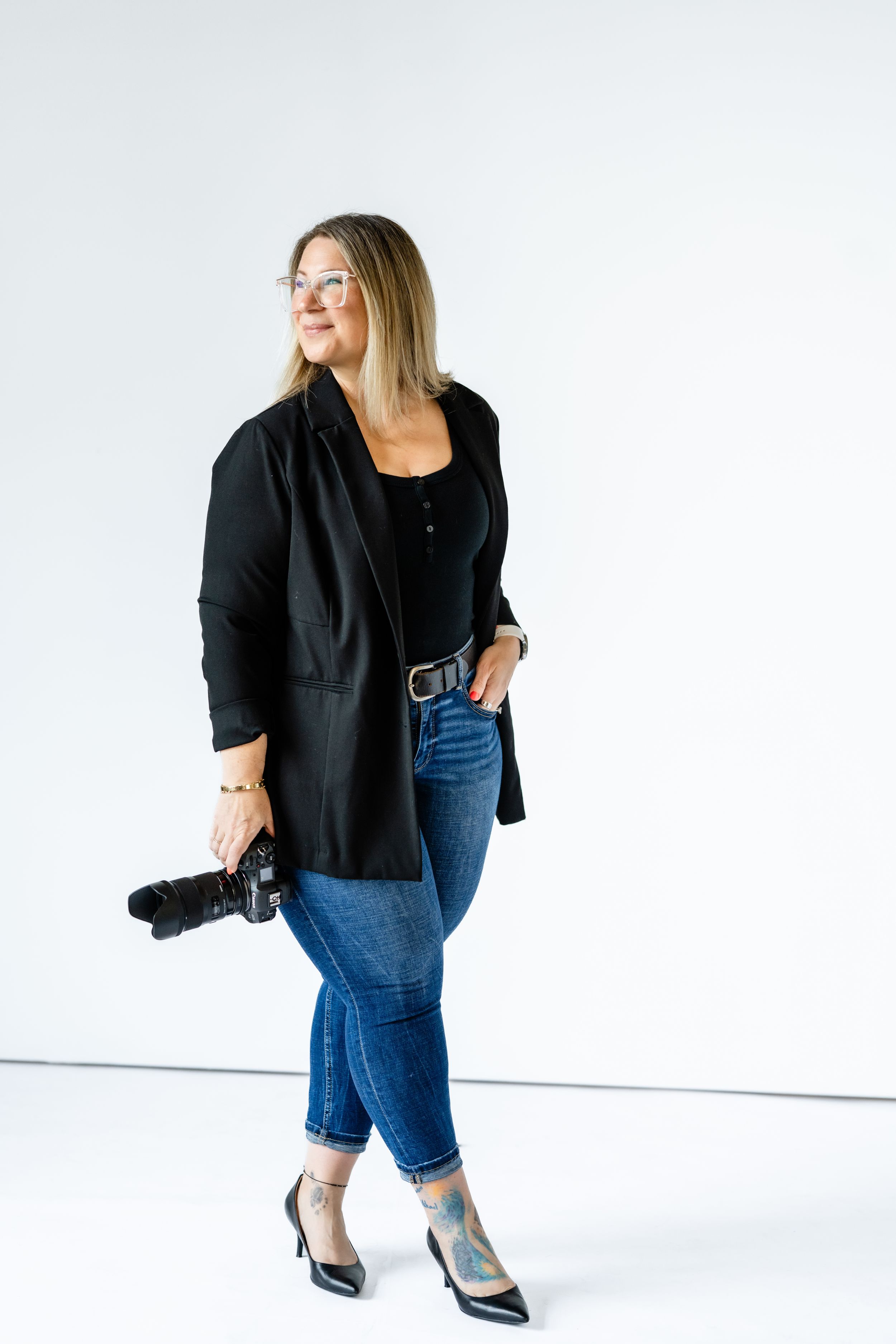 Professional photographer in black blazer and jeans holding camera while posing against white studio background.