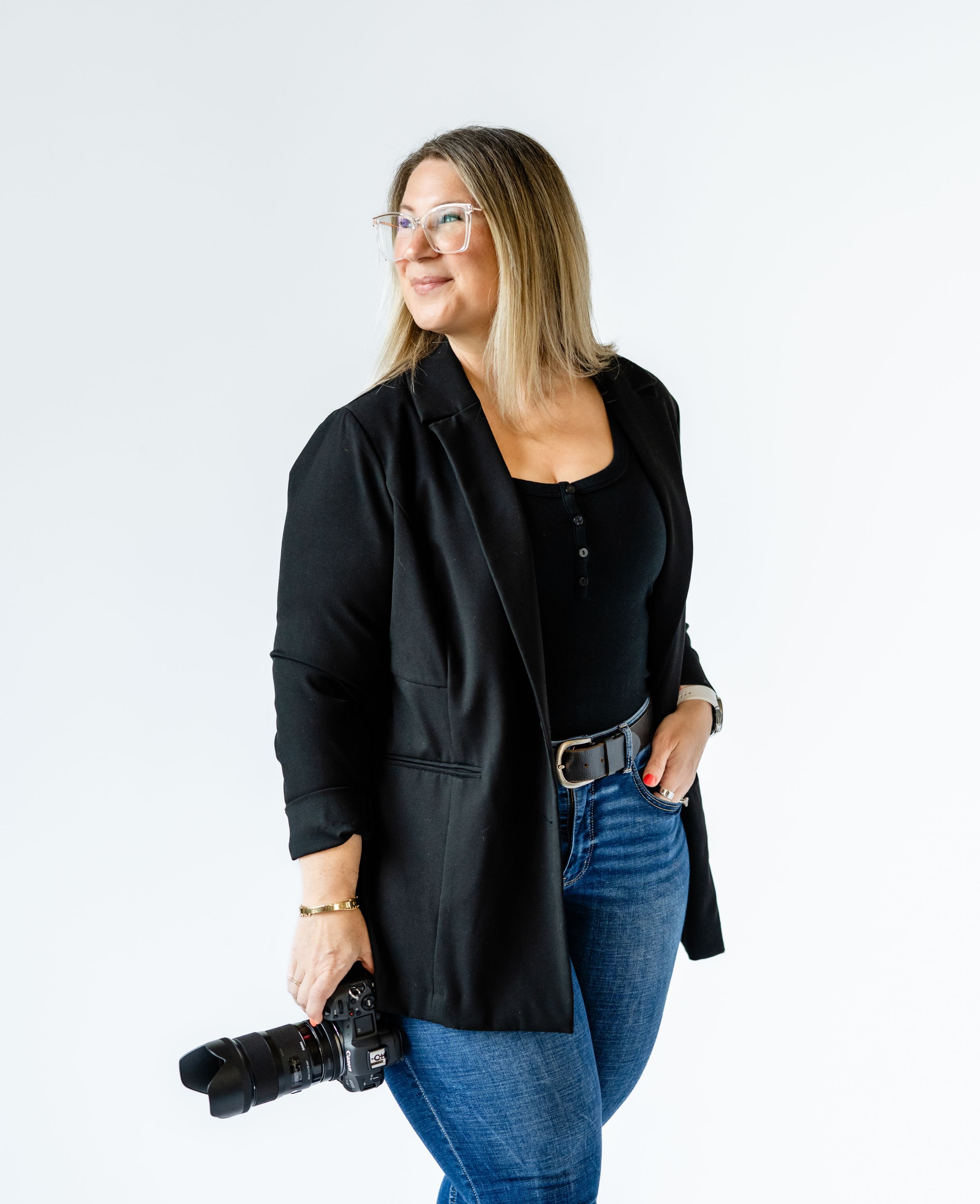 Professional photographer wearing glasses, black blazer and jeans holds camera while standing against white background.