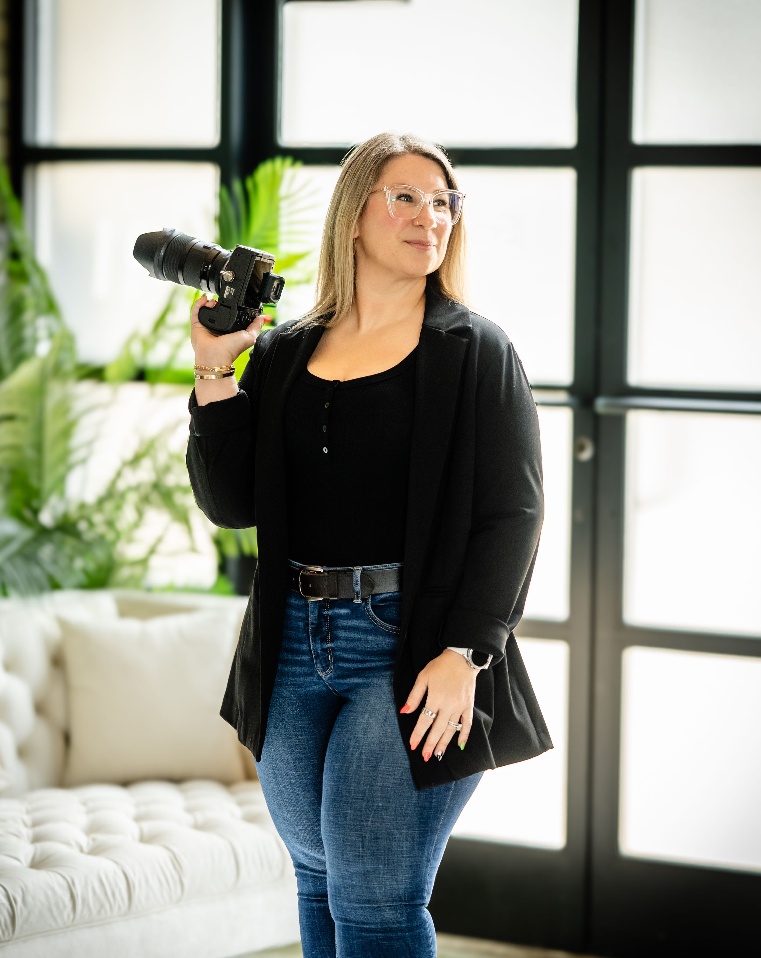 Professional dressed in black blazer and jeans stands confidently by bright window in modern home interior.