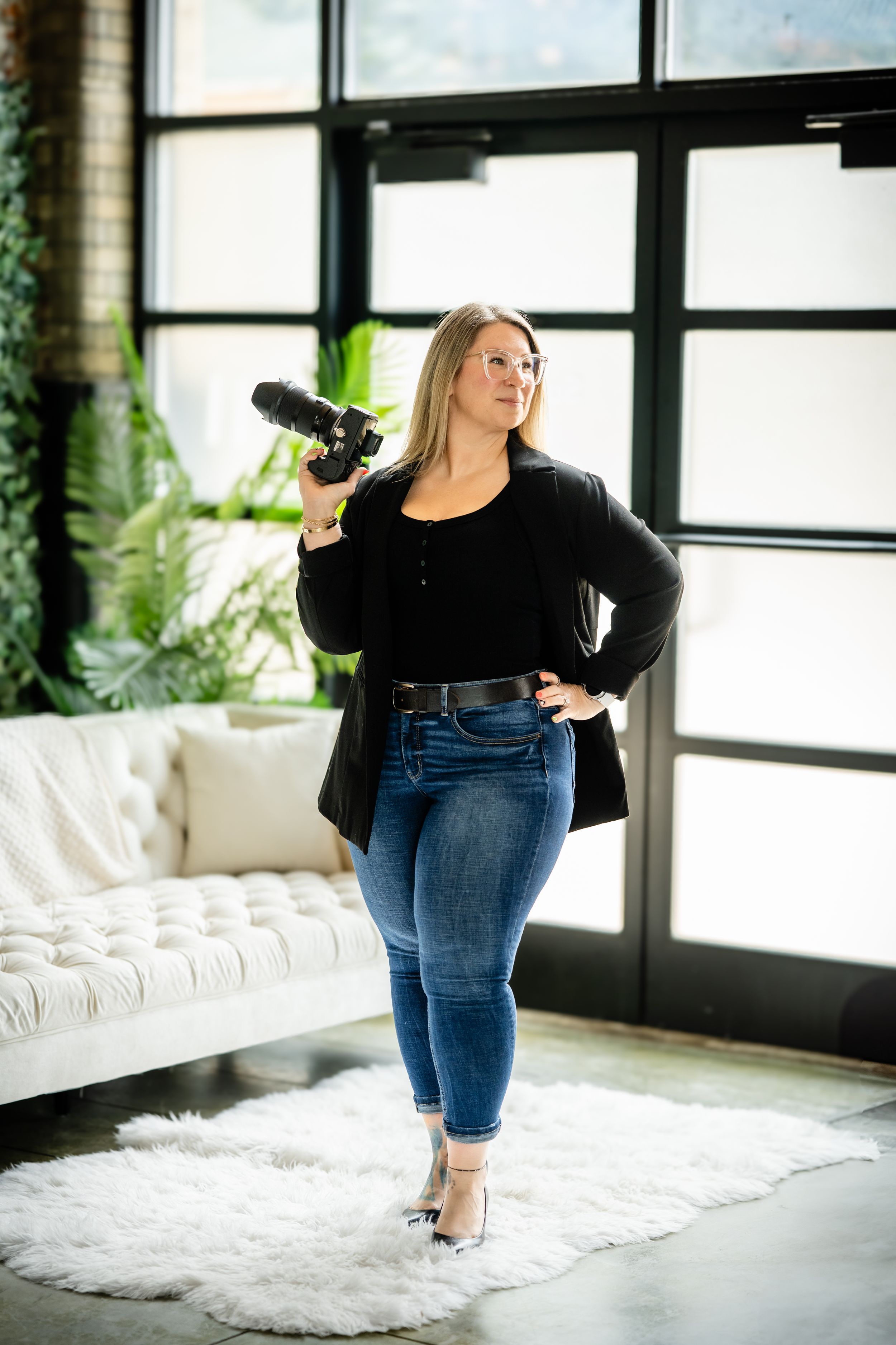 Professional photographer in black top and jeans captures photos while posing on white carpet beside large windows.