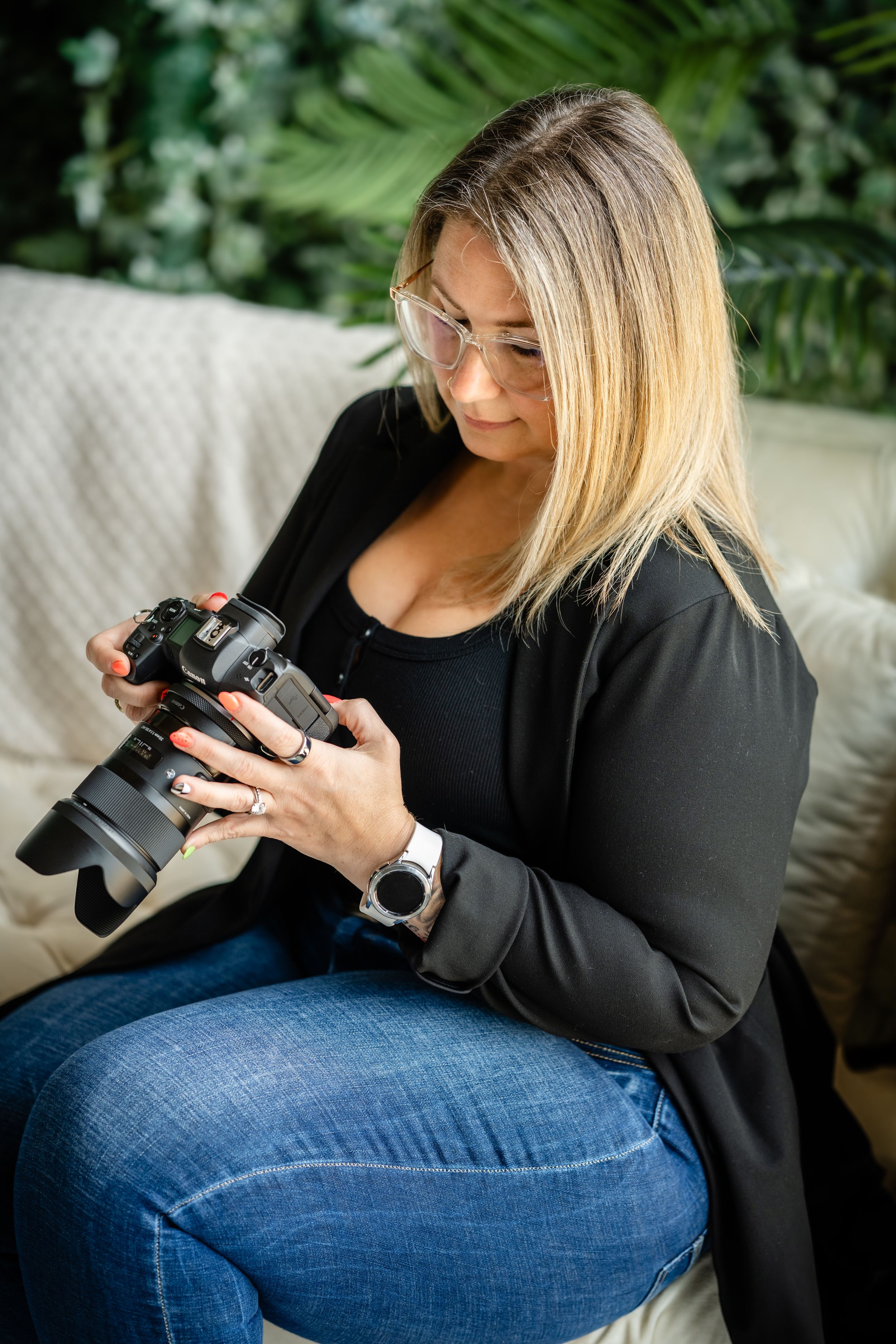 A photographer in a black cardigan and jeans reviews images on a professional camera while sitting on a white couch.