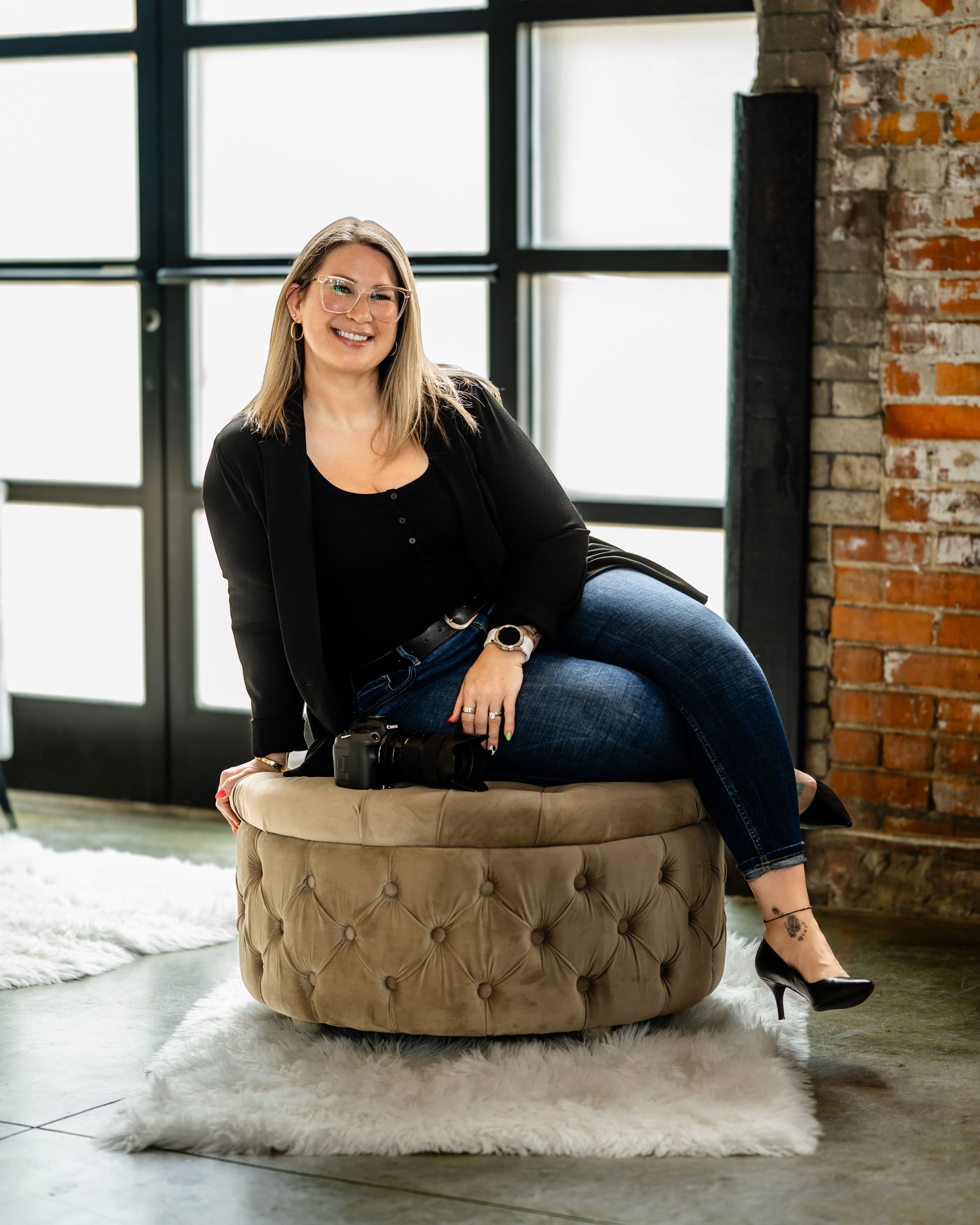 Professional portrait of someone in black clothing casually sitting on a round tufted ottoman in an industrial loft space.
