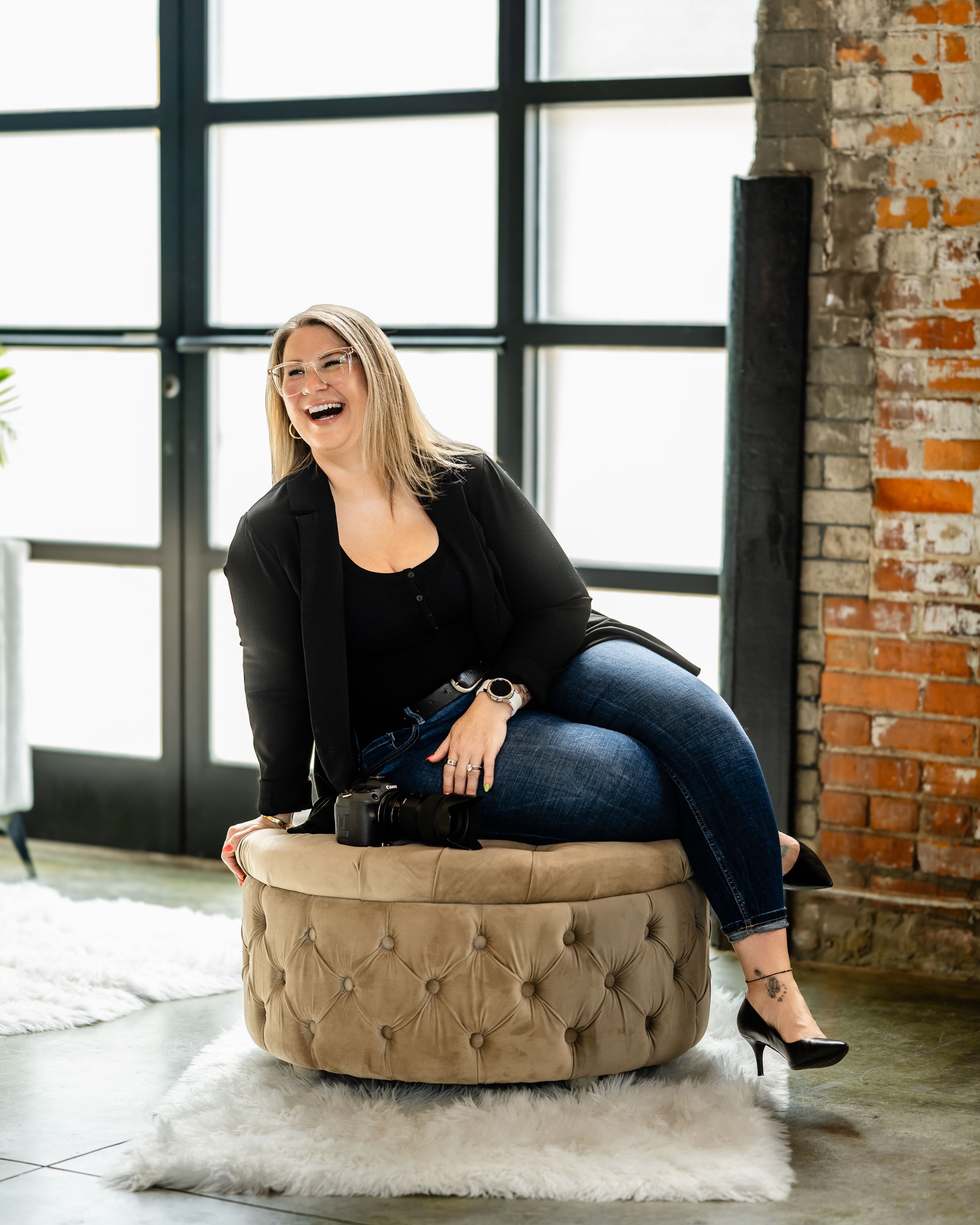Person sitting on a round tufted beige ottoman in an industrial loft space with exposed brick walls and large windows.