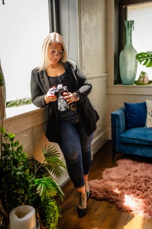 Photographer reviewing images on a digital camera near window with blue velvet chair and indoor plants in cozy home setting.