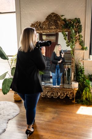 Person in black sweater and jeans looks at reflection in an ornate gold mirror surrounded by plants in sunlit room.