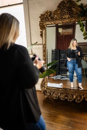 Person taking a selfie in front of an ornate gold-framed floor mirror on hardwood floors with white walls.