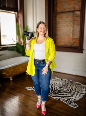 A photographer in a bright yellow blazer, white top and jeans holds a camera while posing on a hardwood floor with a zebra rug.