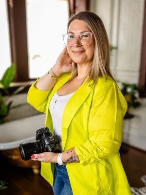 Professional photographer in a bright yellow blazer smiles while holding DSLR camera in sunlit room.