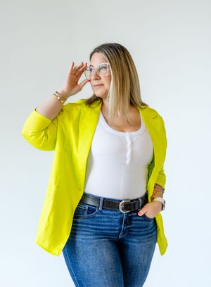 Person wearing a bright yellow blazer with white tank top and jeans poses against white background.