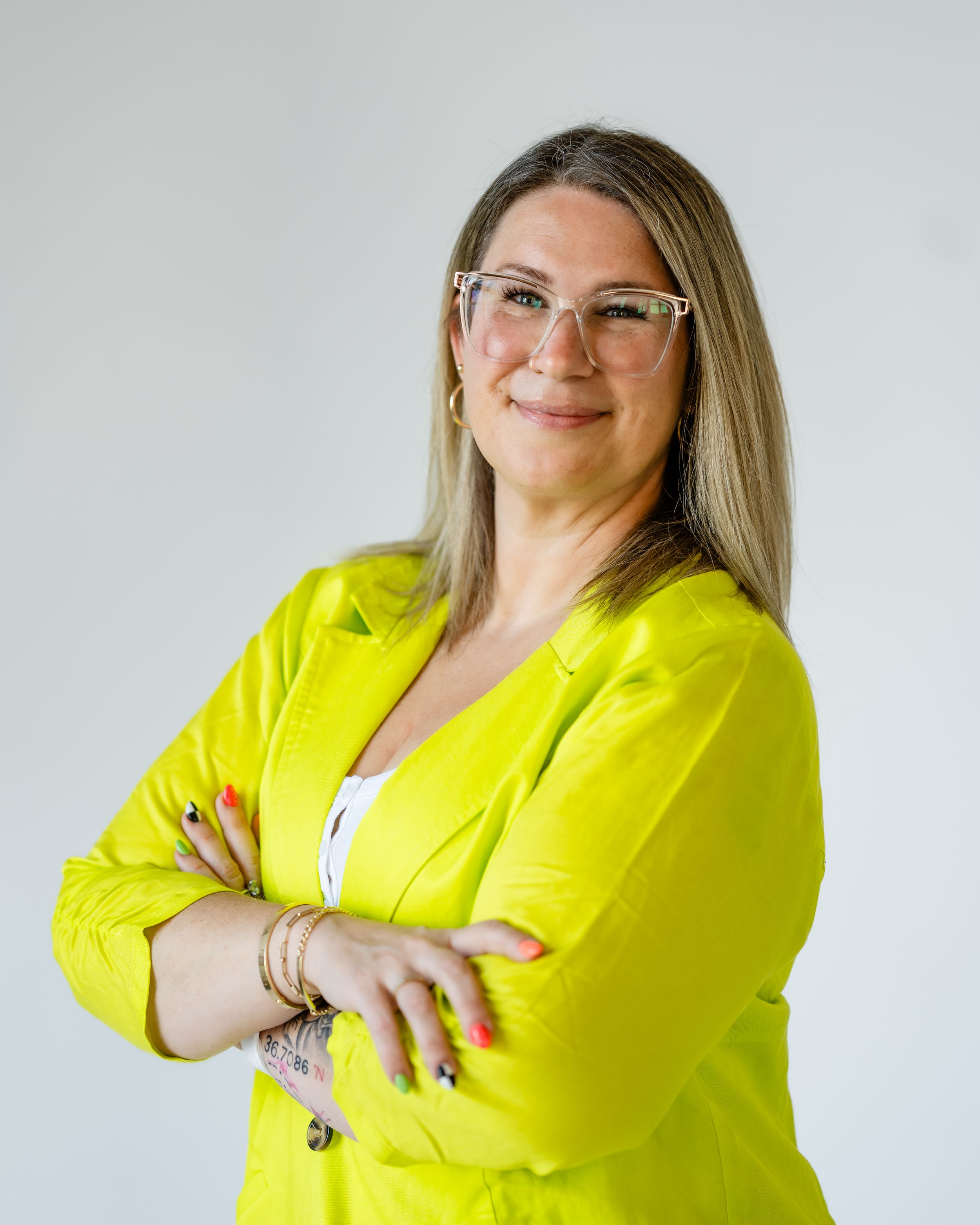Professional portrait of someone in a bright yellow blazer and glasses against a light background, arms crossed.