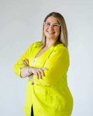 Professional portrait of someone wearing a bright yellow blazer standing with crossed arms against a white background.
